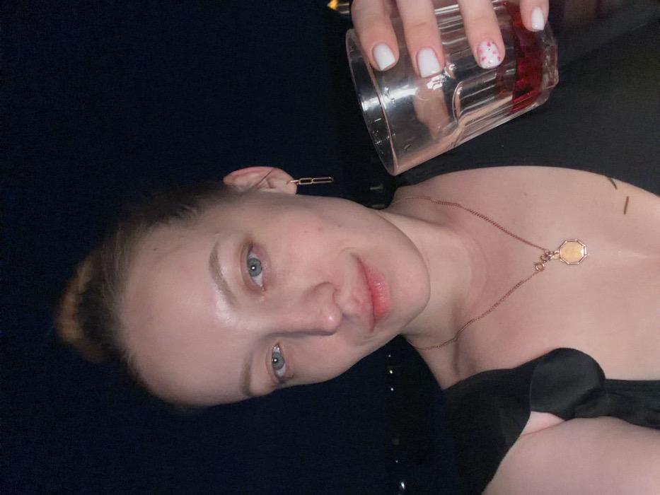 A portrait-style image captures a fair-skinned female subject, appearing to be in her 20s or 30s, looking directly at the camera with a slight smile. Her light-colored hair is styled in a bun. She has light blue eyes. The subject wears a dark-colored top, a gold-toned chain link earring on her right ear (viewer's left), and a gold-toned necklace featuring an octagonal pendant. Her right hand, with nails painted white and the ring finger adorned with red speckles, holds a clear, faceted glass containing a dark red liquid. The background is uniformly dark and out of focus, indicating an outdoor nighttime setting with distant, indistinct lights. This scene is located in Shipulino, Russia.