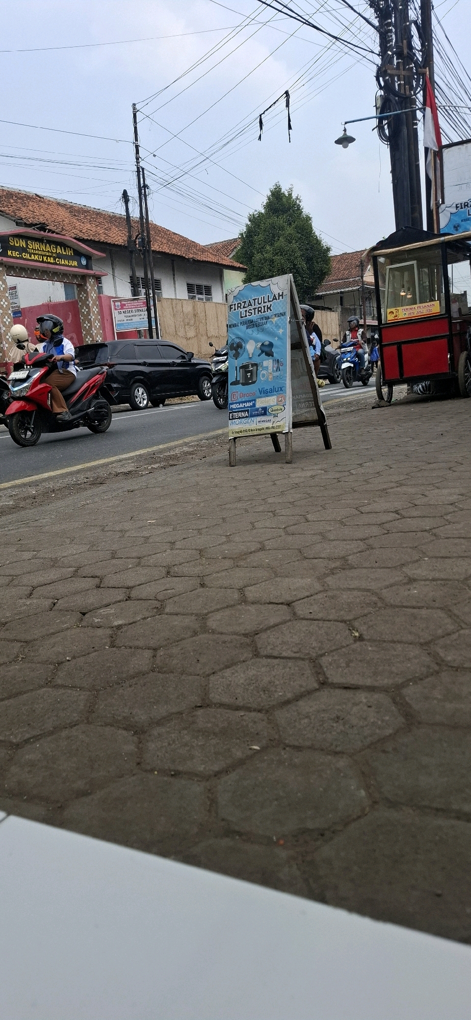 The image depicts a street scene in Sirnagalih, Indonesia, under an overcast sky. In the foreground, a textured, hexagonal-patterned gray sidewalk occupies the bottom portion of the frame. To the right, a red and silver food cart is parked on the sidewalk, featuring a yellow sign with red text that reads "TERIMA PESANAN" (Take Orders) and a phone number.
Further back, a blue A-frame sign with white text is prominently displayed on the sidewalk. The sign advertises "FIRZATULLAH LISTRIK" and lists various electrical equipment brands such as "Braco," "Visalux," "Megaman," "Ploline," "Eterna," "Virbieci," and "Morgen," "Winn Gas." The sign also includes small illustrations of electrical items like light bulbs, a fan, and a rice cooker, along with contact information.
Behind the sign, a black SUV is parked on the right side of the road. To the left of the SUV, a red scooter is being ridden by a person wearing a helmet and a blue and white shirt. Another scooter is visible further back on the left. On the left side of the street, a building with a red tiled roof and white walls is visible, with an entrance gate and a sign that reads "SDN SIRNAGALIIH" (Sirnagalih Elementary School) and "TERAKREDITASI (A)" (Accredited A). The road itself appears to be asphalt, with a yellow line marking the edge. Power lines are visible strung across the sky, supported by utility poles, one of which has an Indonesian flag attached. A green tree stands behind the food cart and to the left of the utility pole. The overall impression is a typical roadside scene in a residential or commercial area.