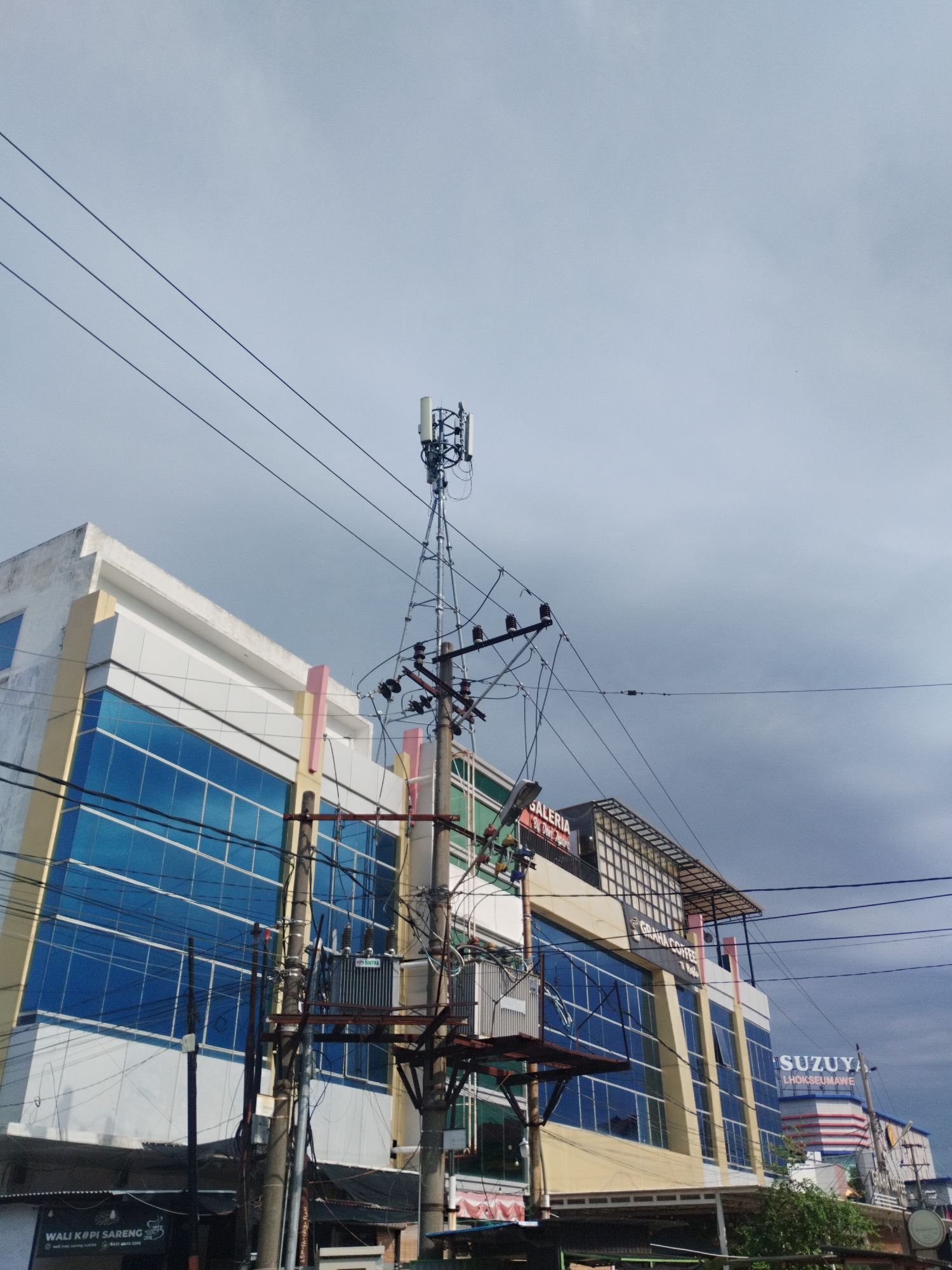 The image shows a view of a street in Lhokseumawe, Indonesia, dominated by a cell tower and a multi-story building. The cell tower, positioned in the center of the frame, is a tall metal structure equipped with antennas. Utility poles with multiple wires and electrical transformers are also prominent features of the scene, suggesting an urban or semi-urban setting with significant electrical infrastructure.
To the left of the cell tower, a modern building with a facade of blue glass windows and cream-colored accents is visible. The building appears to be a commercial establishment, possibly a store or office complex, with signage that is partially visible. To the right of the cell tower, another building is seen with a prominent "SUZUYA" sign and "LHOKSEUMAWE" written below it, identifying the location more precisely. This building also features a contemporary architectural style with glass elements.
The sky is overcast with dark clouds, indicating a cloudy or potentially rainy day. The foreground shows a glimpse of street-level activity with what appears to be shop fronts, including one with a sign that reads "WALI KOPI SARENG". The overall impression is a typical urban streetscape in Indonesia, with a mix of modern and functional infrastructure. There are no people clearly visible in the image, and the focus is on the architectural elements and infrastructure of the street.