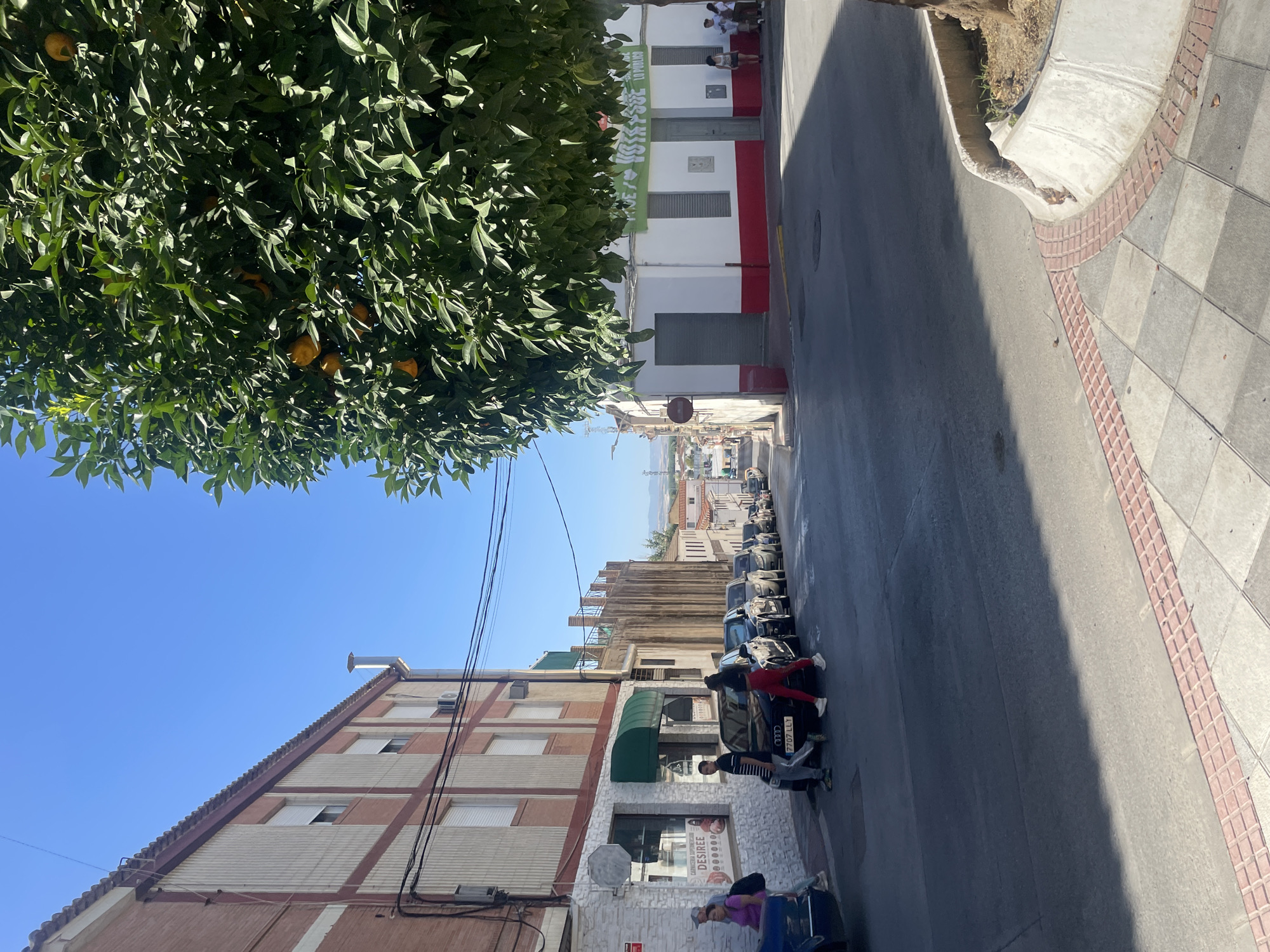 The image captures a street scene in Atarfe, Spain, bathed in bright sunlight. On the left, a multi-story building with a red-tiled roof and brick and cream-colored facade lines the street. Several windows with closed shutters are visible. A store with a white facade and a green awning is situated at the base of this building, displaying text that includes "CARNICERIA SUPERMERCADO DESIREE".
A row of parked cars stretches along the right side of the street, which appears to be sloping downhill. Pedestrians are visible crossing the street in the foreground. One person in a purple shirt and jeans is walking with a backpack, while another in a striped t-shirt and jeans is crossing with two individuals in athletic wear, one in a black top and red pants, and another in a white t-shirt and dark pants.
To the right of the street, an orange tree laden with fruit partially obscures the view of buildings. One building features a white facade with a red base and a green banner displaying text. Another building further down the street has a similar white facade with a red base. The background shows more buildings and a hazy landscape under a clear blue sky. The sidewalk in the foreground is paved with checkered gray and white tiles bordered by red brick accents. A concrete planter with some vegetation is also present.