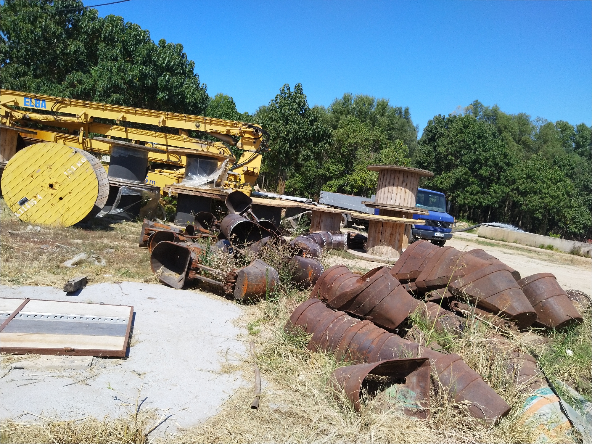 The image depicts an outdoor scene in Valchedram, Bulgaria, featuring industrial equipment and debris. In the foreground and midground, several large, rusty metal containers or drums are scattered on dry grass and concrete. These containers appear to be old and weathered, some are open, revealing their hollow interior. Behind these, a large yellow construction crane is visible, with its arm extended. A large wooden cable spool, painted yellow, is positioned on the left side of the crane. Further in the background, a blue Mercedes-Benz van is parked. The scene is set against a backdrop of lush green trees under a clear blue sky. The overall impression is one of a storage or disposal area for construction and industrial materials.