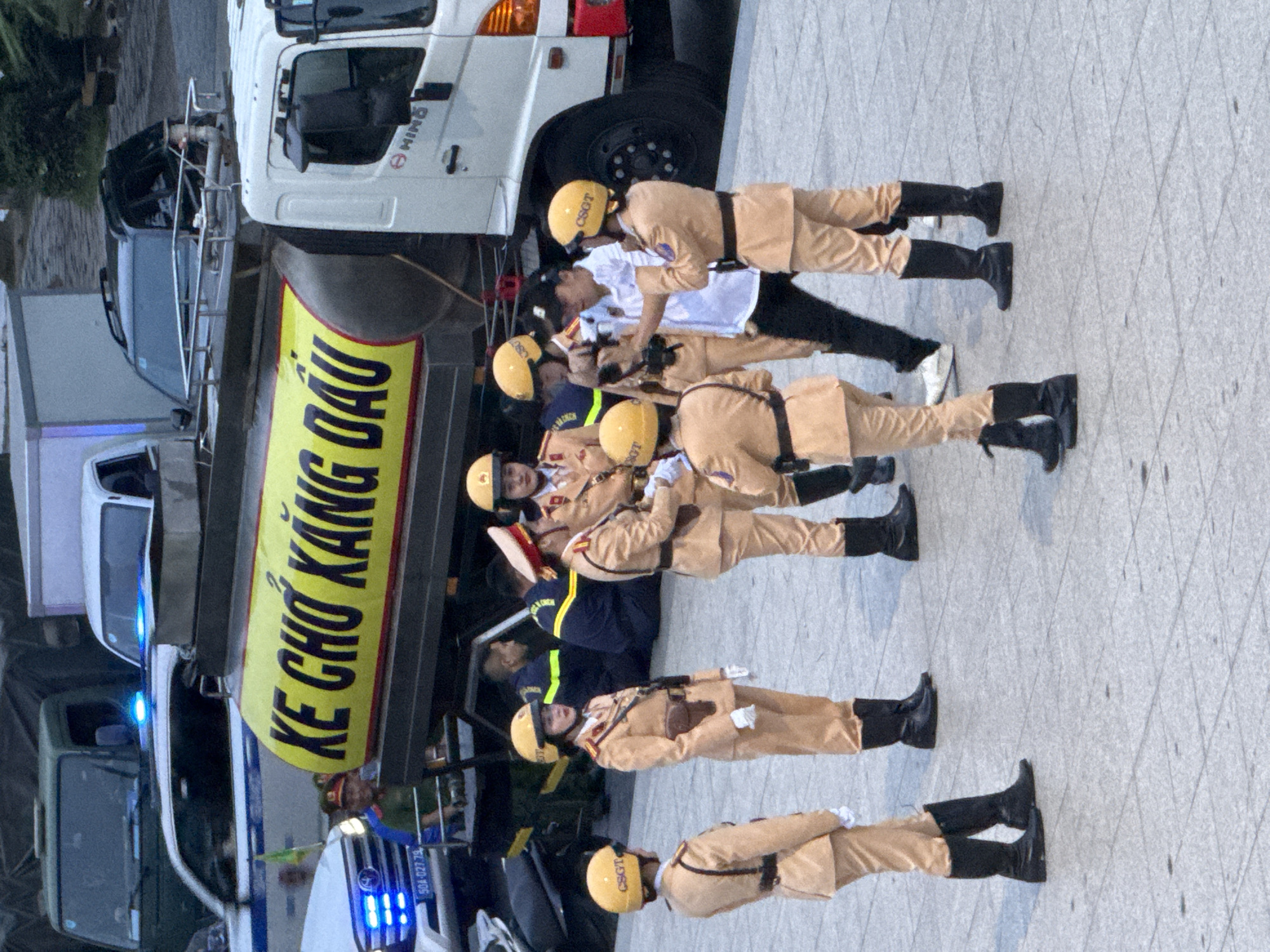 In Da Lat, Vietnam, on a paved plaza, a group of individuals in uniform stand near several vehicles. A large white tank truck with the yellow text "KE CHONG KANG DAU" painted on its side is parked, potentially for a public service event or demonstration. Several officers, identified by their tan uniforms and yellow helmets labeled "CSGT," are positioned in a line, facing towards the camera. One officer in the foreground is looking left, holding their hands in front. Another officer, slightly behind and to the right, faces the camera directly. A man in a white shirt and black pants is in the center of the group, holding what appears to be a camera or recording device and interacting with the officers. Behind the tank truck, a white pickup truck with flashing blue lights is visible, and further back, a dark green truck is also present. The scene appears to be under daylight conditions, casting subtle shadows on the ground.
