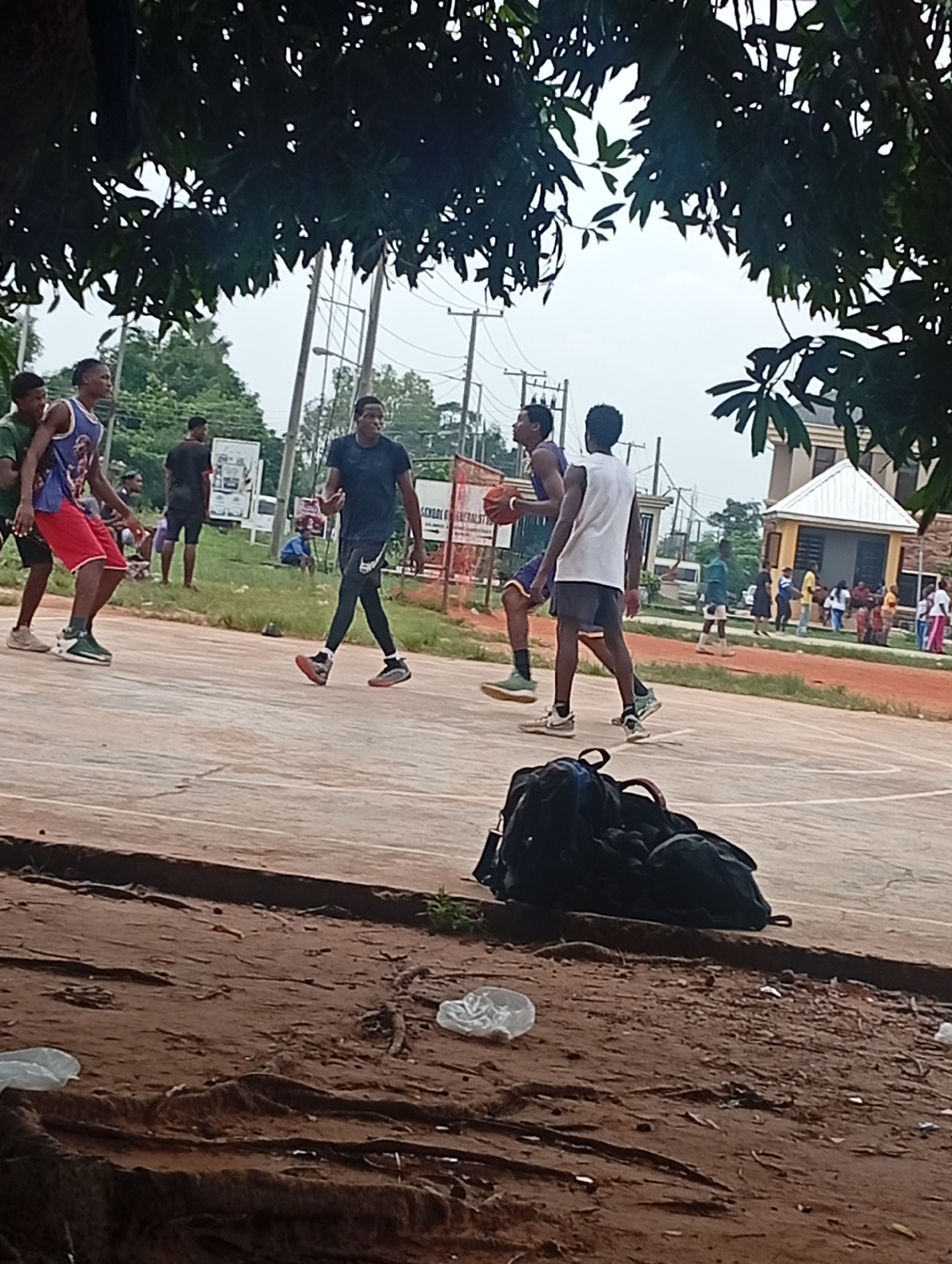 In Ukukwa, Nigeria, a group of young men are engaged in a lively game of basketball on an outdoor court. The scene is set beneath the shade of lush green trees, with a cloudy sky overhead suggesting a potentially overcast day. In the foreground, a pile of dark backpacks rests on the reddish-brown dirt bordering the court.
The main subjects are four young men actively playing basketball. One individual in a blue and white jersey with red shorts is dribbling the ball, while another in a purple and yellow jersey, holding the ball, appears to be passing or shooting. Two other players, one in a black t-shirt and black leggings, and another in a white t-shirt and dark shorts, are actively defending or moving on the court.
In the background, several other individuals are gathered, observing the game or socializing. A small, light-colored building with a distinctive white roof stands to the right, possibly a structure on the sports facility grounds. The presence of utility poles and overhead wires indicates an urban or semi-urban setting. A white board with visible text is placed near the basketball hoop on the left side of the court. The overall atmosphere appears casual and energetic, typical of a friendly pickup basketball game.