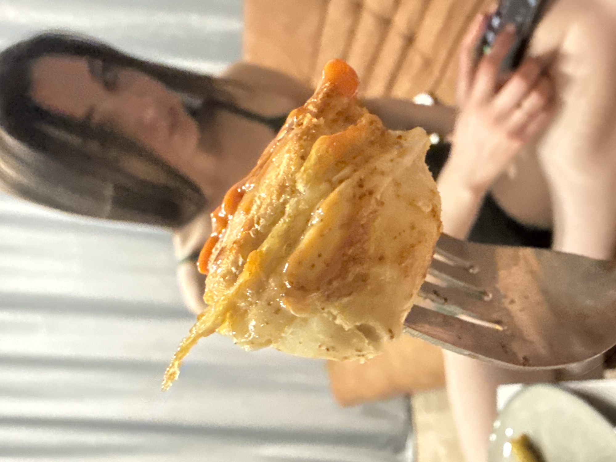 A close-up, shallow depth-of-field photograph captures a piece of food, likely a pancake, crepe, or pastry, impaled on a silver fork in the foreground. The food item is soft and golden-brown, generously coated in a glistening, viscous sauce, possibly caramel or syrup, which appears to be dripping slightly from its edges.
In the blurred background, a person with long, dark hair is partially visible on the left, looking downwards. Their face is out of focus, making emotions indistinguishable. On the right, their left hand is holding a dark object, which appears to be a smartphone. Part of their bare shoulder or arm is also visible. The background suggests an indoor setting, with hints of wooden textures and what might be another plate below the fork. The overall activity depicted is a person about to eat or in the process of eating. The lighting is soft, consistent with indoor conditions. No text is visible in the image.