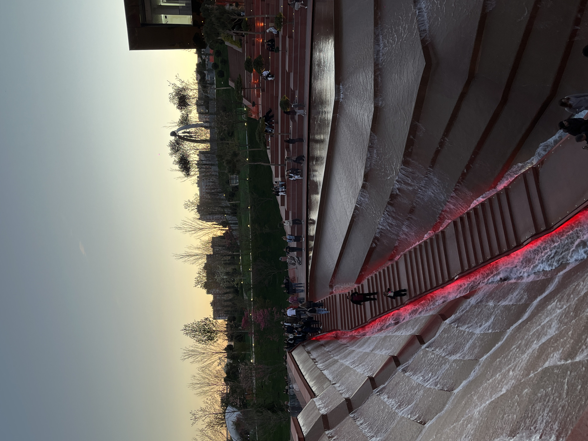 This image, taken in Krasnodar, Russia, depicts an outdoor public space, likely a park or plaza, at sunset or during the golden hour. The dominant feature is a large, multi-tiered water cascade or fountain constructed from reddish-brown material. Water flows over each tier, creating a dynamic visual effect. A notable detail is a striking red LED light strip illuminating the edge where the water meets the adjacent wide steps, casting a vibrant glow on the cascading water.
Numerous people are present throughout the scene. Many are seen walking or sitting on the wide, matching reddish-brown steps that run parallel to the water feature, enjoying the evening. In the park area on the left side of the image, there is green grass, numerous trees (many appearing bare, suggesting late winter or early spring), and a distinct abstract, light-colored ring-like sculpture. Distant buildings are visible in the background, indicating an urban setting.
The sky displays a soft gradient, transitioning from a warm, yellowish hue near the horizon to a lighter blue-grey overhead, characteristic of a clear evening. The overall atmosphere appears peaceful and leisurely, with people casually enjoying the architectural and natural elements of the park. There is no visible text in the media file.