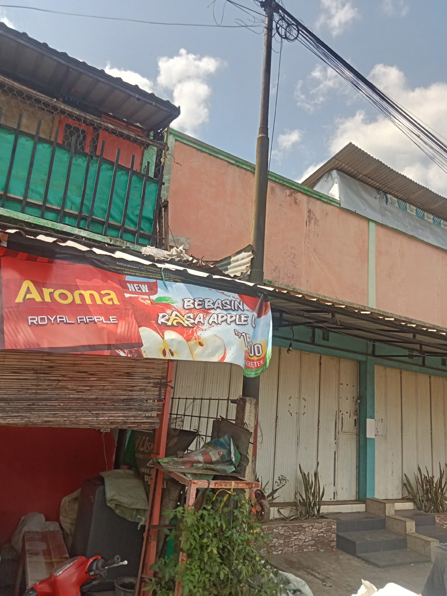 A roadside view in Sawangan, Indonesia, shows a building with a prominent banner advertising "Aroma Royal Apple" flavored cigarettes. The building has a peach-colored facade and a corrugated metal roof. A utility pole with numerous wires stands in front of the building. To the left of the building, a red scooter is parked near a red wall and various stacked items. In front of the building, roll-up metal doors are visible, and a small set of stairs leads up to the entrance. Several potted snake plants are placed near the stairs. The sky above is partly cloudy.