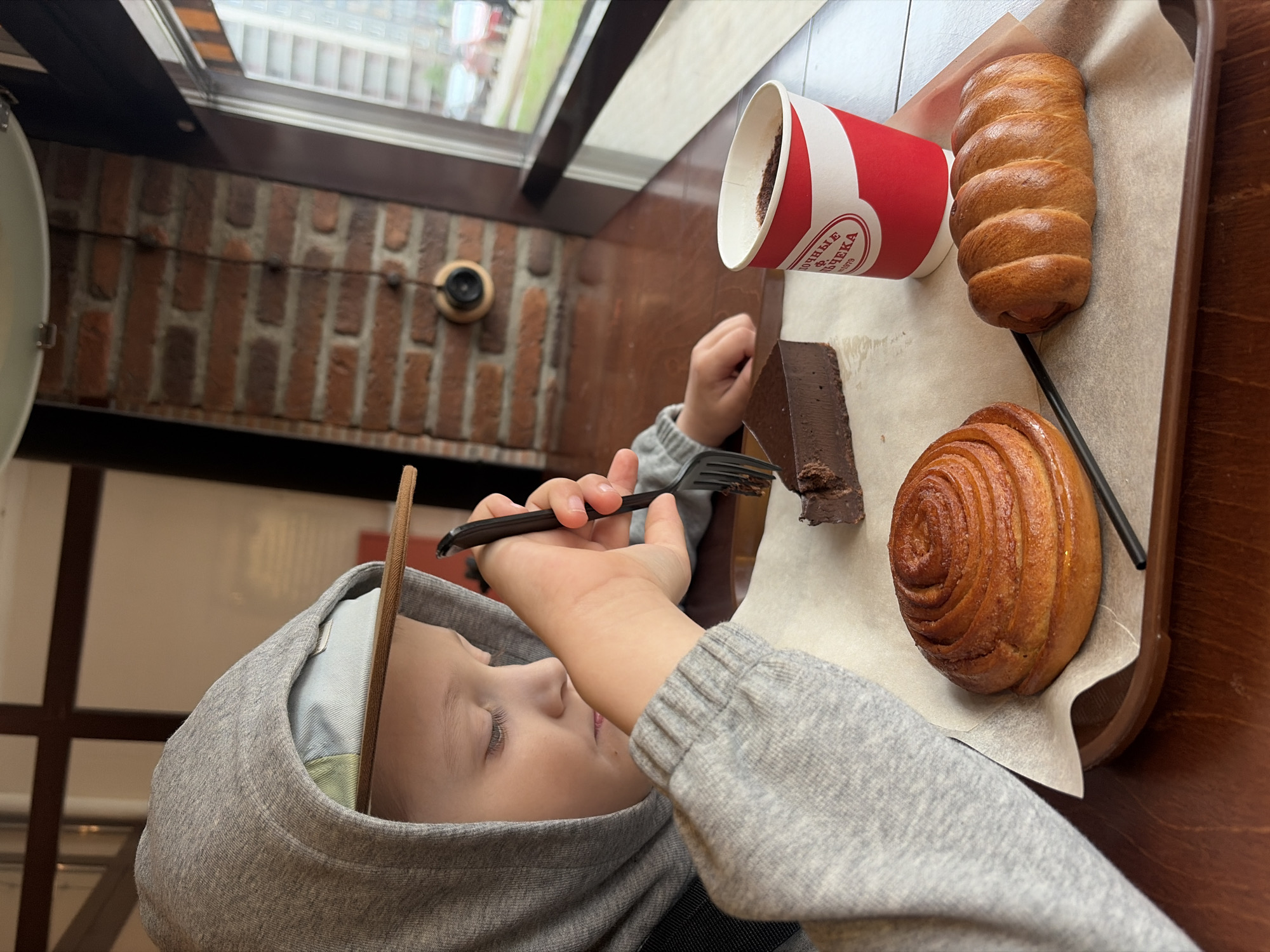 A young child in a gray hooded sweatshirt sits at a wooden table in what appears to be a cafe. The child is holding a fork and eating a slice of chocolate cake from a tray containing other pastries: a cinnamon roll and a filled bun. A red and white paper cup stands next to the cake. The location is Saint Petersburg, Russia, indicated by the Russian inscription on the paper cup, which translates to "Bulochka Factory" (булочные фабрики) and the year "1870." The background features a brick wall and a window, suggesting an indoor setting.