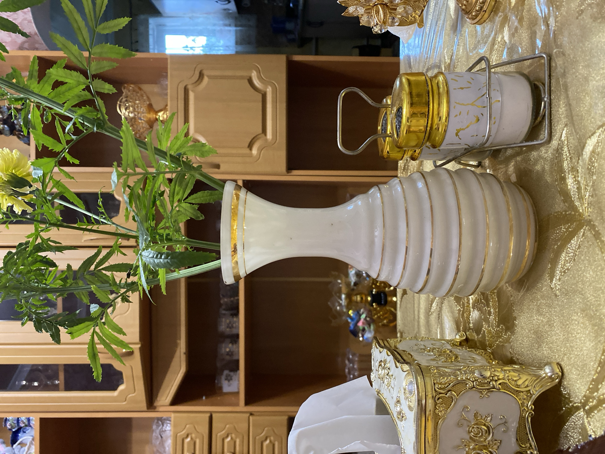 The image depicts an indoor scene, likely a dining room or living room, with a focus on a table set with decorative items. The table is covered with a shimmering gold fabric tablecloth. Prominently featured on the table is a white ceramic vase with gold trim, containing a tall green plant with serrated leaves and yellow flowers. To the left of the vase is an ornate, rectangular tissue box with gold detailing and what appears to be floral motifs. To the right of the vase are two small glass shakers with gold lids, held in a silver-toned metal caddy.
In the background, a wooden cabinet with multiple shelves and decorative doors is visible. Some shelves display various objects, including what appear to be small decorative items and potentially glasses or containers. A window with sheer curtains is also visible on the far right side of the background. The overall impression is one of an elegantly decorated interior space. There are no people visible in the image. The location is not specified, and cannot be inferred from the image.