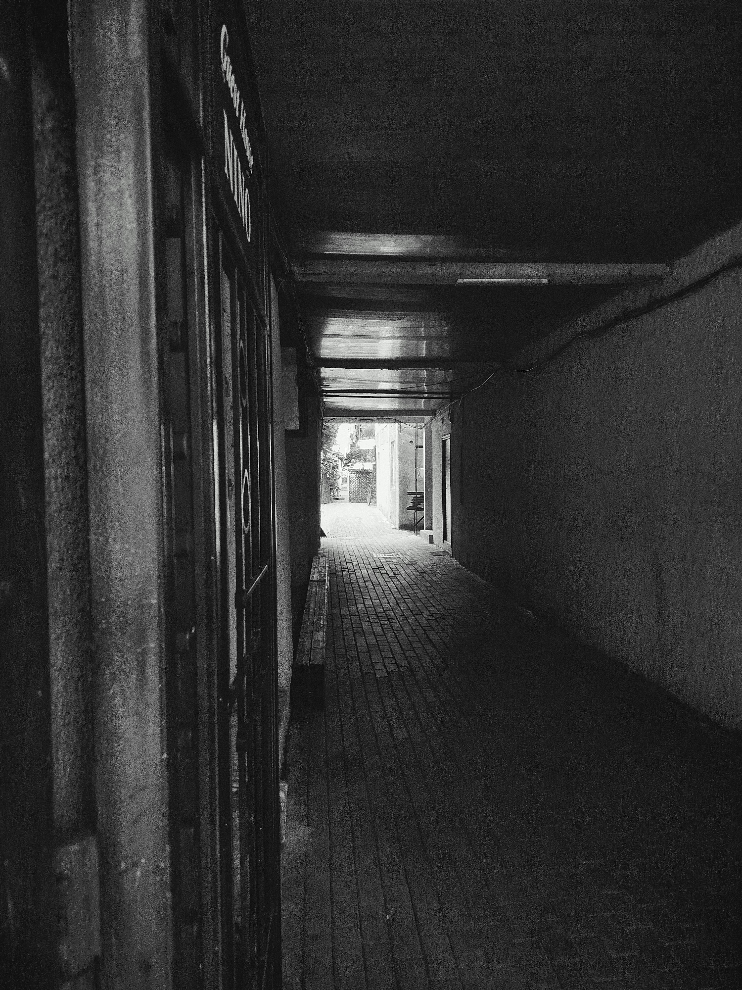 The image depicts a narrow, covered walkway with a brick-paved floor. On the left, a weathered metal gate and a concrete pillar are prominent. A sign attached to the wall above the gate reads "Guest House Nino." The walkway is dimly lit, with light coming from the end, suggesting an outdoor area. The walls on either side are rough textured. A simple wooden bench is visible along the right wall. The overall impression is one of an alleyway or a passage leading into an open space, possibly within the old town of Batumi, Georgia. There are no people visible in the image.
