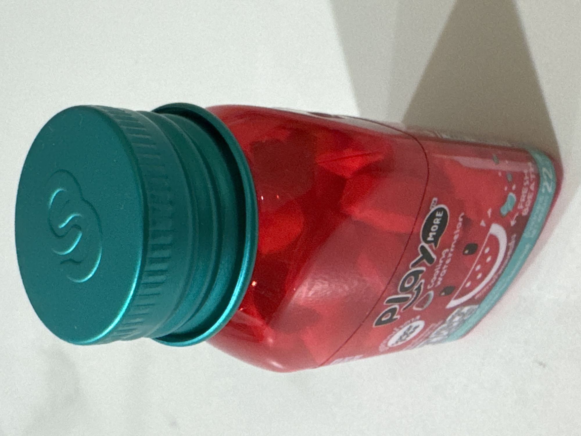 The image shows a close-up, angled shot of a red, translucent bottle with a teal-colored cap. The bottle appears to be made of plastic. The cap has a textured surface and a stylized "S" logo embossed on its top. The bottle's label is partially visible, displaying text that includes "PLAY MORE" and "Cooling watermelon". There are also graphics of watermelon slices and small dots scattered across the label. A shadow is cast on the white surface behind the bottle, suggesting illumination from a light source. The overall impression is that of a product shot, likely for marketing or product identification purposes. The scene is a simple, clean white background, focusing attention entirely on the product. There are no people or other objects present in the frame. The lighting suggests an indoor setting, possibly a studio or a well-lit room. The time of day and weather are not discernible from the image. The location context "Nha Trang Ward, Vietnam" does not provide any visual cues within the image itself.