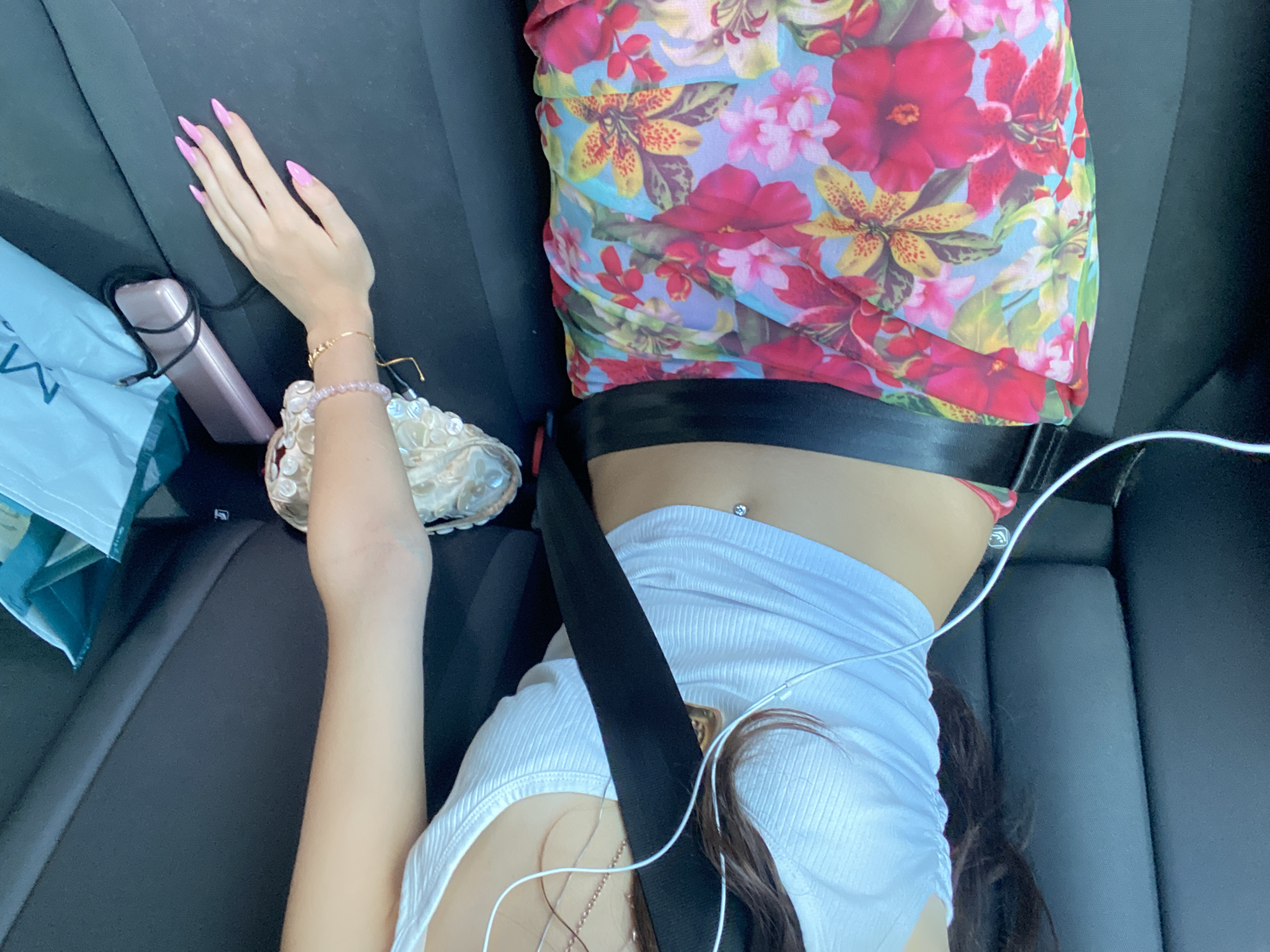 The image shows a person lying down in what appears to be the backseat of a car. The person is wearing a white ribbed tank top and a colorful floral skirt. A black seatbelt is fastened across their torso. White earbuds are plugged into the person's ear, with the wire running down. A belly button piercing is visible. The person's hand, adorned with bright pink stiletto nails and bracelets, rests on the car seat. A pink electronic device and a light blue bag are also present in the car. The scene is dimly lit, suggesting it might be inside a car with the windows tinted or at night. There is no discernible information in the image that would place this scene specifically in Mole Valley, United Kingdom.