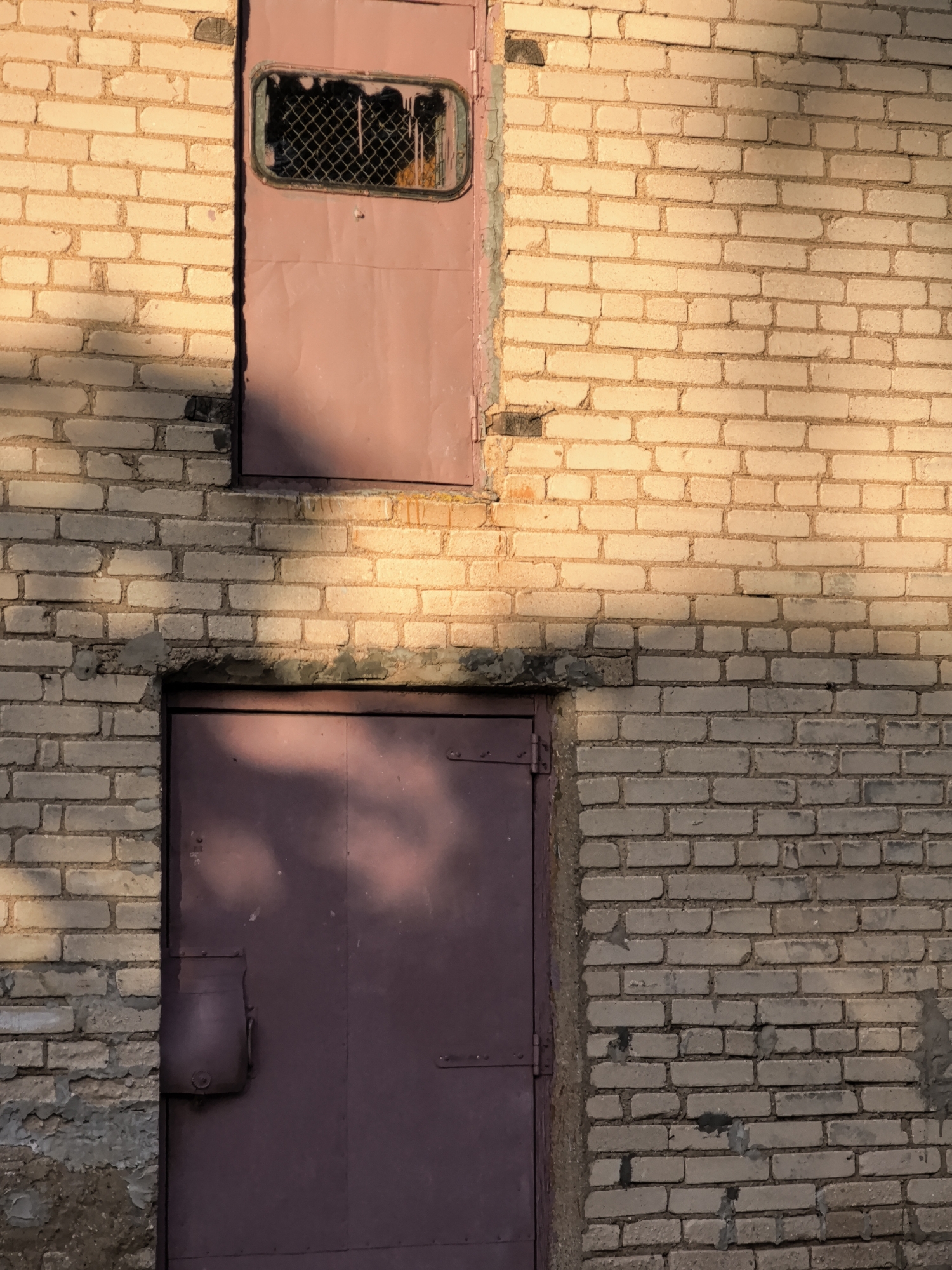 An exterior view of a light-colored brick wall with two doors. The upper door is smaller, made of metal, and painted a muted rose color. It has a rectangular window with a metal grate that is broken and shows a dark interior. The lower door is larger, also metal and painted the same rose color, and appears to be a double door. It has a boxy metal handle mechanism on the left side. The brick wall shows signs of age with some mortar missing and discoloration. Sunlight casts shadows across the wall, highlighting the texture of the bricks and the surface of the doors.
