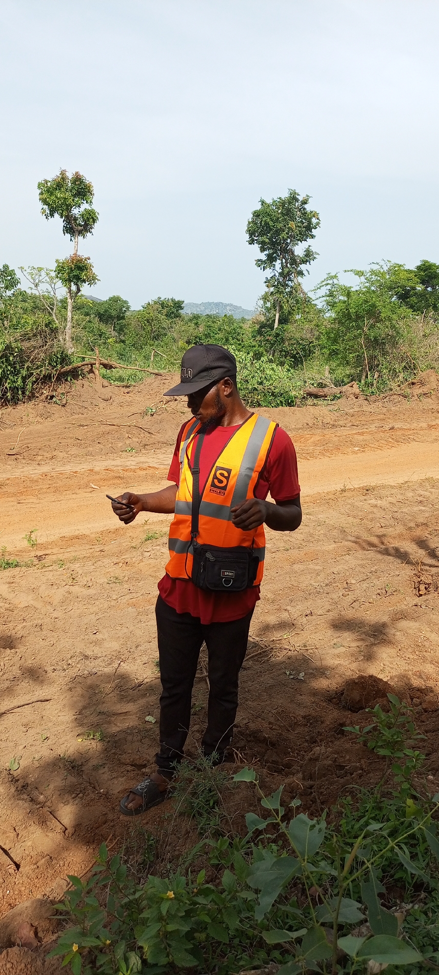 A man stands outdoors in a cleared, earthy area, likely in Otunade, Nigeria. He is of Black ethnicity, wearing a black baseball cap with "OLD" visible on the front, a red short-sleeved t-shirt, and an orange high-visibility vest with reflective stripes. On the left side of his vest, a logo with a large "S" and "SWALBYS" underneath is visible. He also wears black pants and black open-toed sandals. A black waist bag with "SPORT" written on it is worn across his chest. He is holding a small dark object, likely a smartphone, in his left hand and is looking down at it with a focused expression.
The setting appears to be a construction site or recently cleared land, characterized by reddish-brown, uneven dirt with visible tracks or disturbances. Patches of green vegetation and small plants are scattered on the ground, particularly in the foreground. In the mid-ground and background, numerous trees and dense foliage are present, with distant hills visible under a pale blue, slightly hazy sky. The lighting suggests it is daytime, possibly mid-morning or early afternoon, with shadows cast by the man indicating the sun's position.