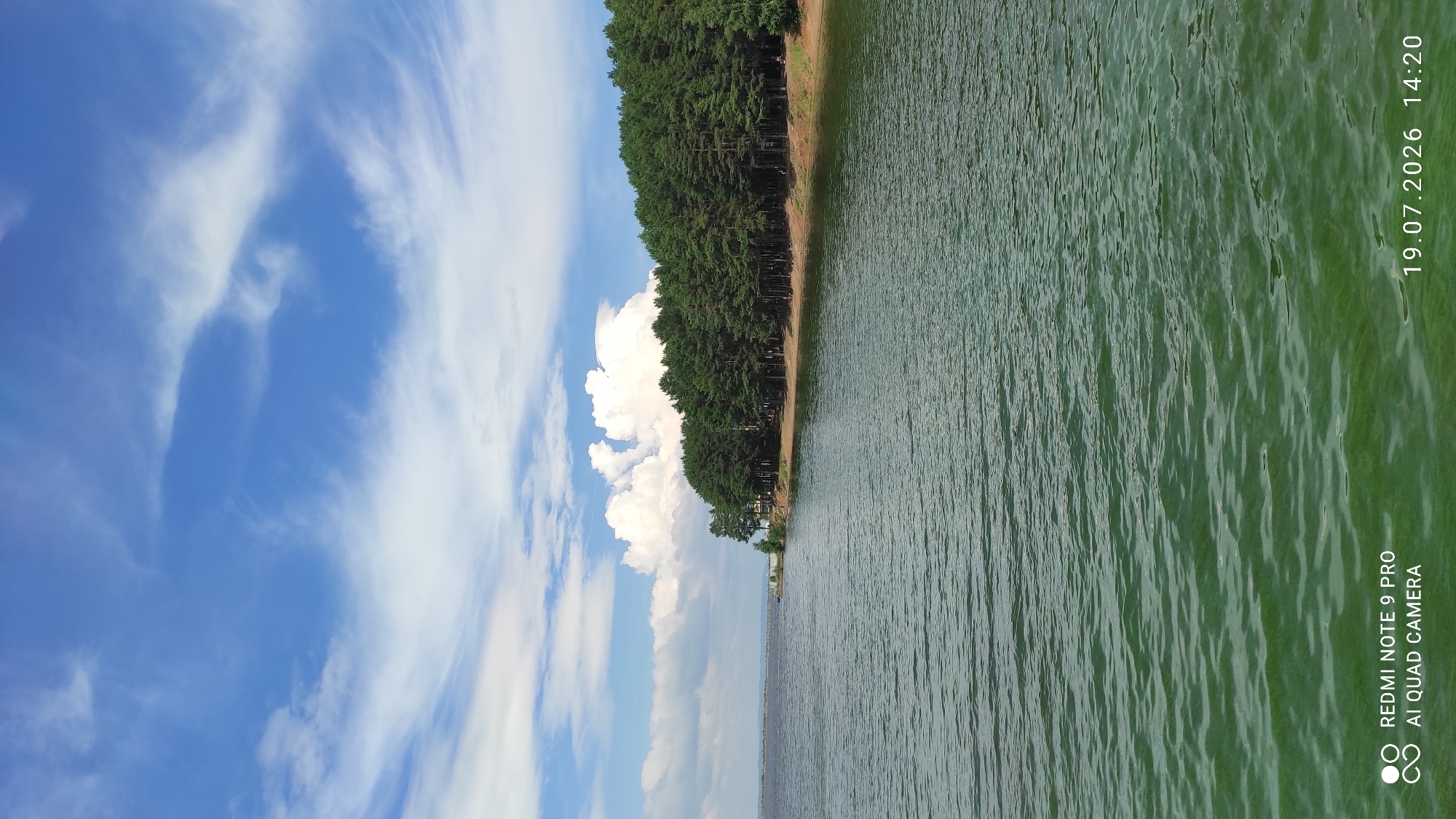 This image depicts a body of water, likely a reservoir, with a shoreline lined by a dense forest of coniferous trees. The water is a muted green color, with visible ripples on its surface indicating a slight breeze. The sky above is a vibrant blue, dotted with white, wispy clouds. The trees are tall and appear to be pine or fir, forming a thick, green canopy along the edge of the water. The shoreline itself is a sandy or earthy brown. The overall scene conveys a sense of natural tranquility. The bottom left corner of the image displays text indicating it was taken with a "REDMI NOTE 9 PRO AI QUAD CAMERA" on "19.07.2026 14:20". It is plausible this location is near the city of Chaikovsky, Russia, given the typical landscape of the region which includes large water bodies and forested areas.