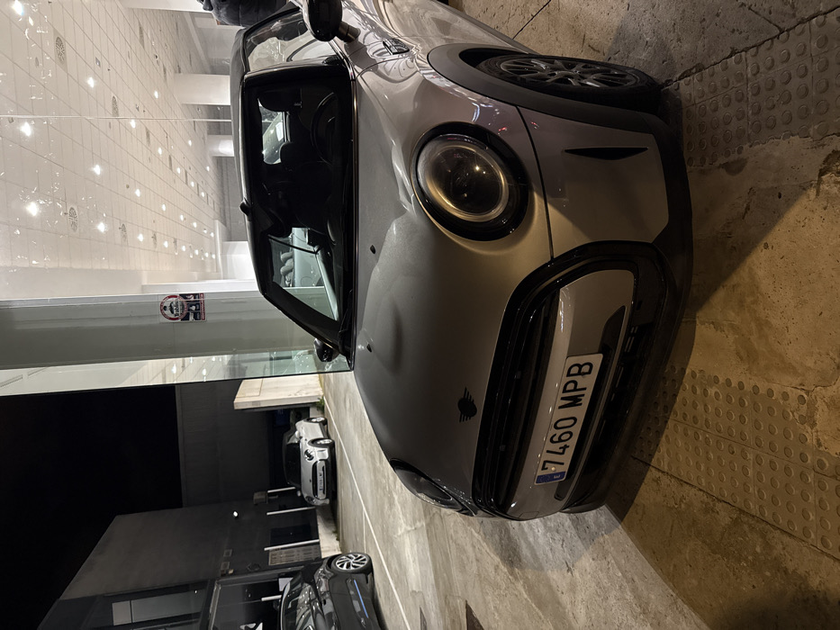 The scene depicts a silver Mini Cooper convertible, license plate '7460 MPB', parked at night on a light-colored concrete surface with raised tactile paving elements, within the Polígon Industrial De Son Castello, Spain. The vehicle is angled slightly towards the left, with its front fascia, left headlight, and a portion of the hood prominently featured. The car's soft top appears lowered, and the interior is dark. To the right of the Mini, a large glass window reflects numerous interior overhead lights from a brightly lit commercial space, likely a showroom or dealership. A red and white "Prohibido el Paso" (No Entry) sign is affixed to a vertical glass column or pillar separating the glass panels. In the background, to the left, a dark grey vehicle is partially visible, parked on a similar concrete surface. Further back, a white Mini Cooper convertible is parked in front of a dark, utilitarian building facade under the night sky. No individuals are visible in the image. The lighting is primarily artificial, originating from the building interior and possibly streetlights off-frame, casting shadows on the ground.