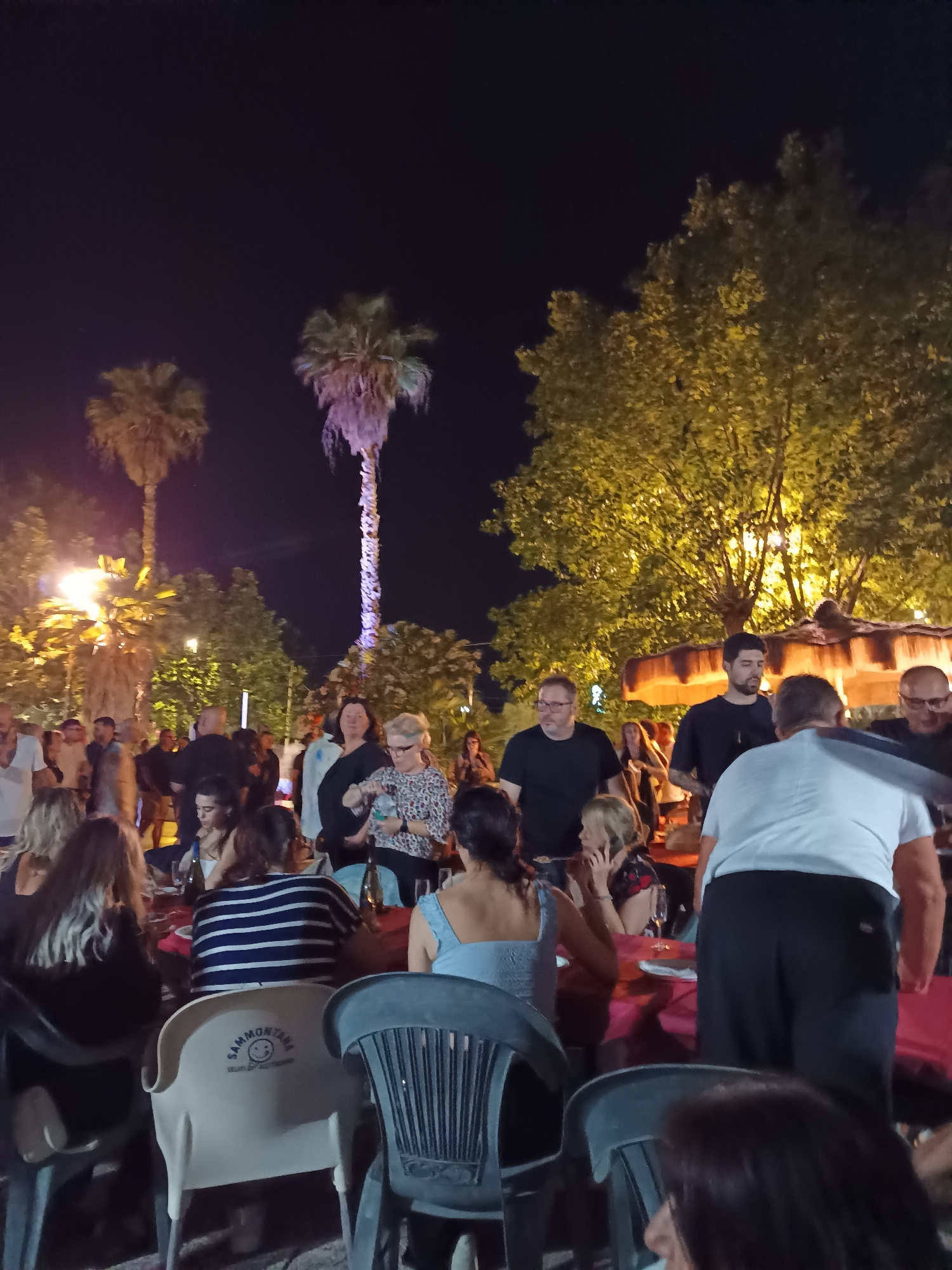 The image depicts an outdoor gathering at night, possibly a restaurant or event space, with numerous people seated and standing around tables. Palm trees are visible in the background, illuminated by artificial lights, and trees with yellow foliage are also present. The scene appears to be lively, with groups of people interacting. One person is holding a bottle of water. The chairs at the tables are a mix of plastic and some with upholstery. A white chair in the foreground has text and a smiley face logo on its backrest. The overall lighting suggests it is nighttime, with the warm glow of lights creating an inviting atmosphere. While the precise location isn't explicitly stated in the image itself, it's described as being in Giulianova, Italy.