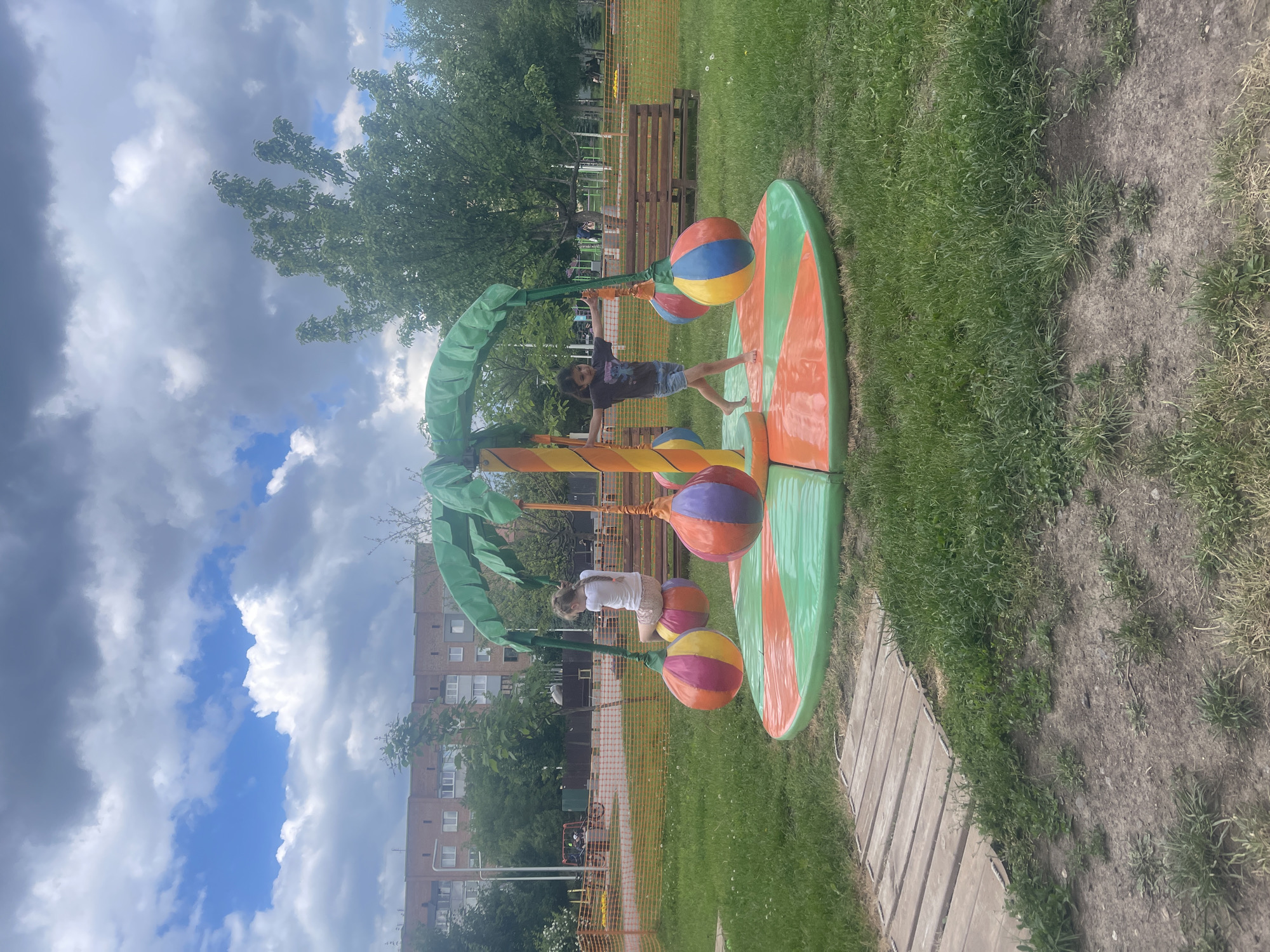 The image depicts an outdoor children's playground in an urban setting. In the foreground, a vibrant, multi-colored playground structure resembling a palm tree is centrally located. Two young girls are interacting with the structure. One girl, wearing a white t-shirt and patterned shorts, sits on a hanging, ball-shaped seat. The other girl, in a dark t-shirt and denim shorts, stands barefoot on the spinning platform, with her arms outstretched. The playground is situated on a patch of green grass, bordered by a wooden pathway on the left and a protective orange net fence in the background. Beyond the fence, wooden benches and trees are visible, and further in the distance, a multi-story brick building stands under a partially cloudy sky. The scene appears to be a bright, possibly late spring or summer day.