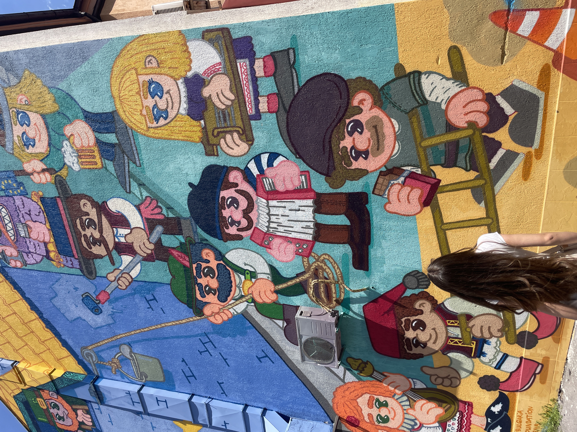 The image is a close-up photograph of a mural painting on a building in Sofia, Bulgaria. The mural features cartoonish figures dressed in what appears to be folk or historical costumes, each engaged in different activities. One figure is holding a ladder, another a string, one is painting a roof, etc. The mural is painted in bright colors. Part of a young woman's head and long brown hair are visible in the lower right corner of the image, near the mural. The building appears to be made of brick, with a section painted yellow, and what appears to be an air conditioning unit visible at the top of the frame. In the lower right corner of the image there is the name "Bulgaria Foundation RMAD". The image has been taken at an angle, making the mural appear slanted. The sky is visible in the top left corner of the frame, indicating it is daytime.