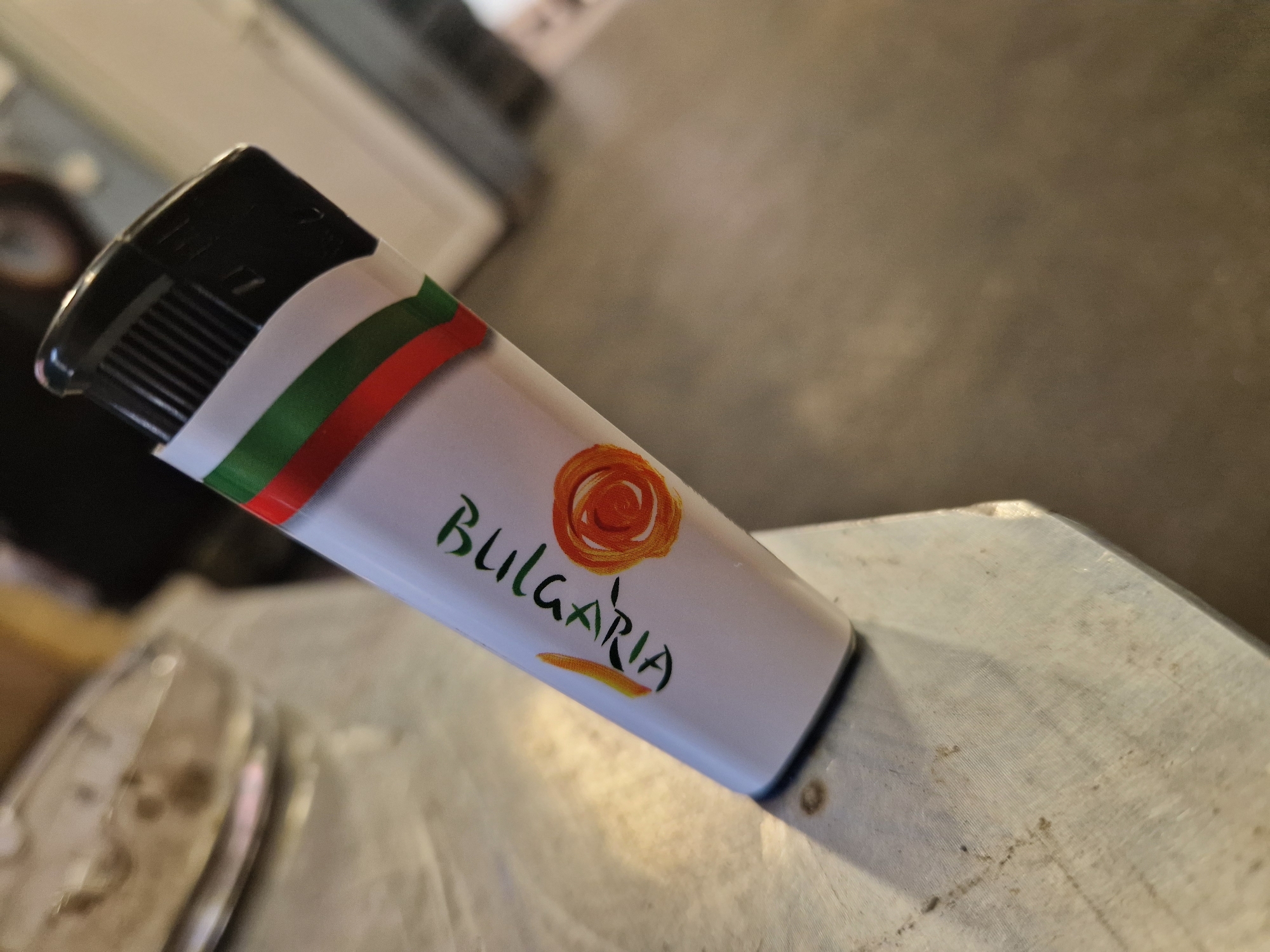 A close-up, slightly angled shot features a white disposable lighter resting on a textured surface. The lighter's top portion is black, with visible grates and mechanisms for ignition. A decorative band encircles the lighter's body, displaying the colors green, white, and red, arranged horizontally from left to right when viewed from the lighter's top. These colors represent the Bulgarian national flag.
Below this colored band, a stylized circular design in shades of orange and yellow, resembling a rose or a swirling sun, is printed. Underneath the design, the word "BULGARIA" is written in a vibrant green, flowing script, with a small orange-yellow brushstroke accent beneath the text.
The lighter rests on a light-colored, possibly metallic or laminate, surface that exhibits some subtle texture and minor discoloration or debris. The background is significantly blurred and out of focus, suggesting an indoor setting. Indistinct shapes and variations in light and shadow are visible in the background, but no specific objects or people are discernible. The overall lighting appears soft and diffused, not indicating a specific time of day. The presence of the Bulgarian flag colors and the word "BULGARIA," coupled with the provided location context, confirms the setting is Sofia, Bulgaria. No activity is depicted; the image is a still life focusing on the souvenir lighter.