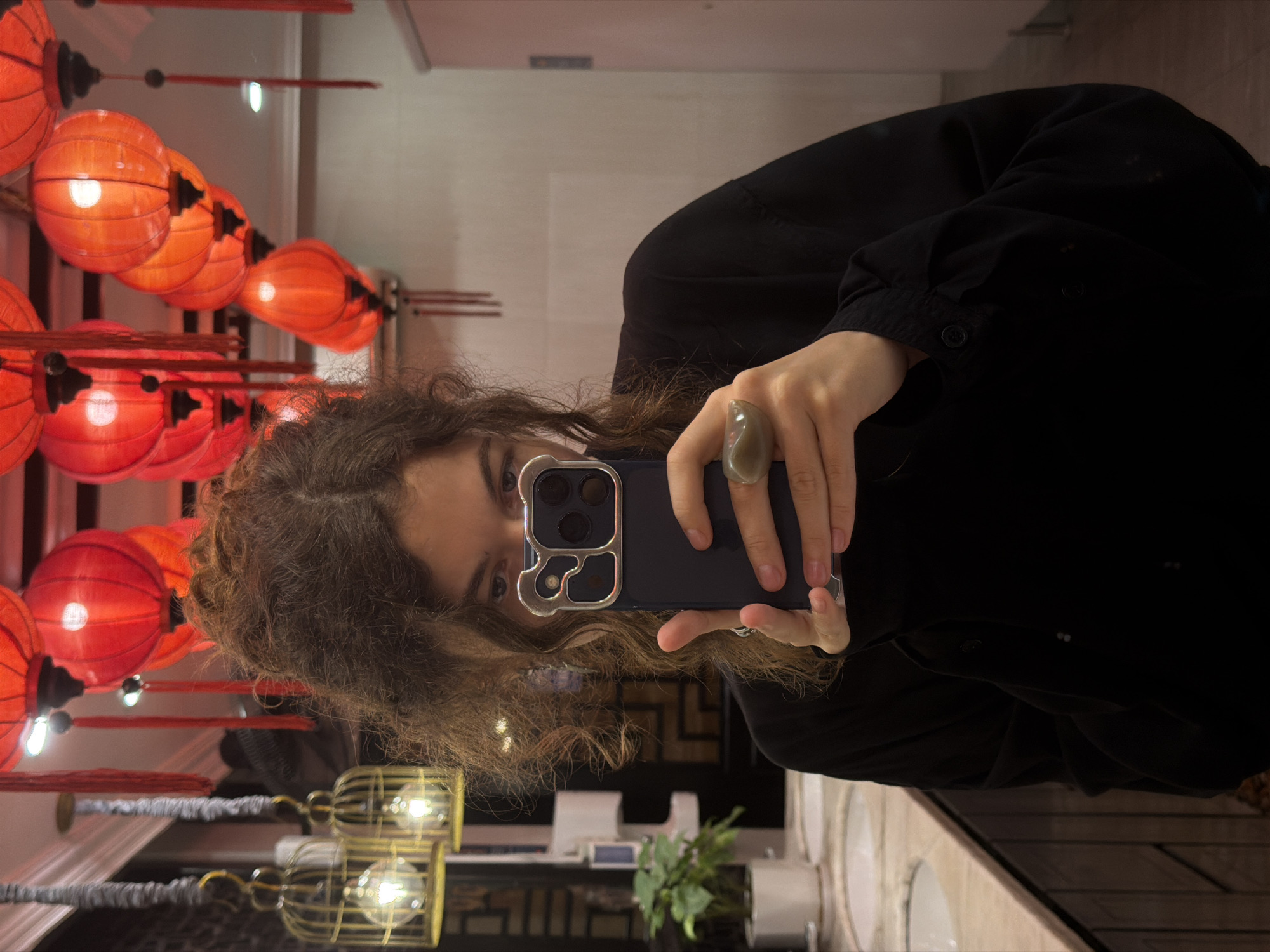 A mirror selfie captures a person with long, curly, dark brown hair and a dark long-sleeved top. The person holds a dark blue smartphone, fitted with a distinctive silver/grey camera protector, in their right hand. A large, translucent light green/grey ring adorns their right index finger, and a white earbud is visible in their left ear. Only their eyes and forehead are clearly reflected in the mirror.
The background, visible in the mirror's reflection, is richly decorated with numerous red and orange Chinese-style lanterns of various shapes and sizes, emitting a warm, soft glow. These lanterns hang from the ceiling, creating an elaborate display. Below these, golden cage-like light fixtures with visible bulbs further illuminate the space.
The setting appears to be a well-decorated interior, likely a public restroom or vanity area, indicated by the long counter with multiple white sinks and a small potted green plant. The vibrant lanterns and ornate lighting suggest an Asian-inspired decor, aligning with the provided context of Đà Nẵng, Vietnam. The lighting implies an indoor environment, possibly during the evening or night. No discernible text is visible.