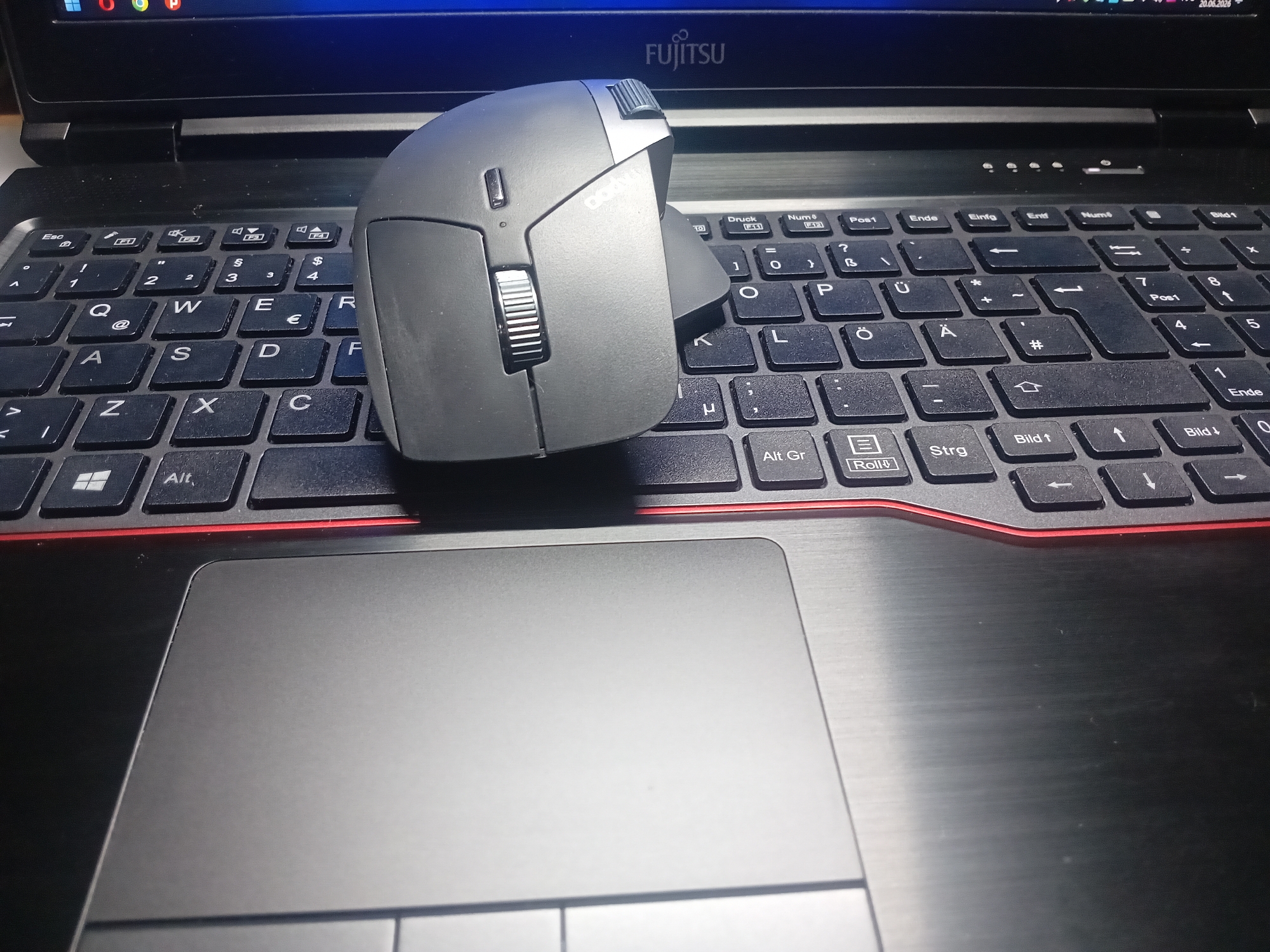 The image shows a close-up, high-angle view of a black laptop with a gray mouse placed on the keyboard. The laptop screen is visible at the top, displaying a portion of a graphical user interface and the word "FUJITSU." The keyboard is a standard QWERTZ layout, with keys such as "Esc," "1," "2," "3," "4," "Q," "W," "E," "R," "A," "S," "D," "F," "Z," "X," "C," and various function keys and modifiers visible. The mouse is ergonomic and gray, with a prominent scroll wheel and several buttons. The red trim around the keyboard area is also visible. The laptop's trackpad is partially visible in the foreground, to the left of the keyboard. The scene appears to be indoors, with artificial lighting. There are no people visible in the image. The location of Bohodukhiv, Ukraine is not indicated by any visual cues in the image.