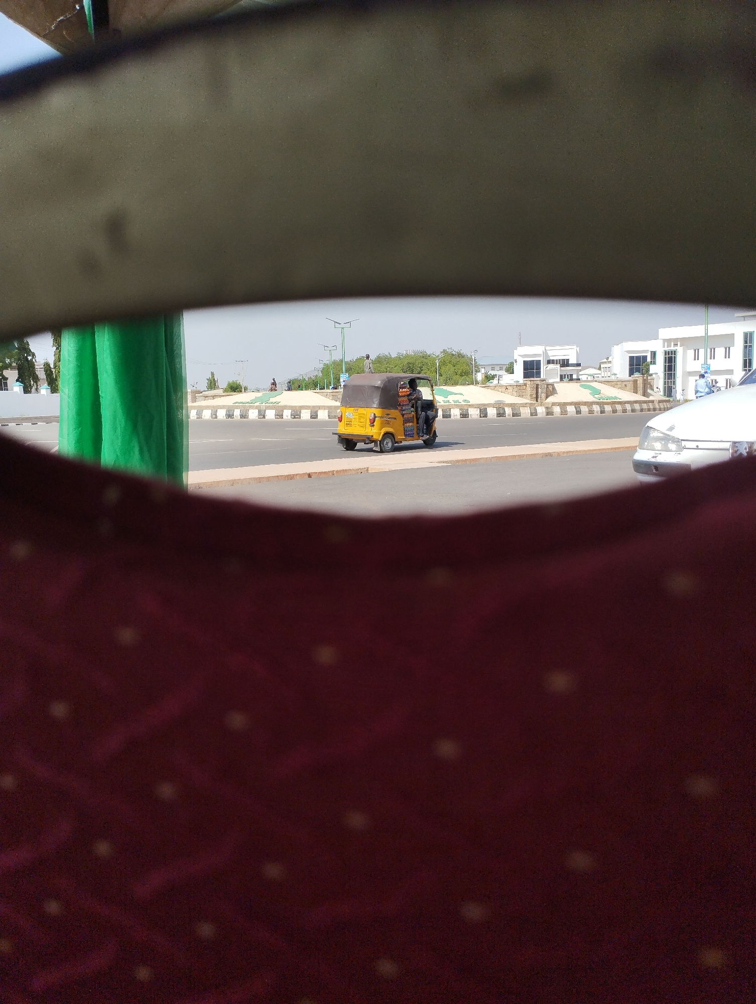 The image, taken from inside a vehicle, shows a yellow tricycle taxi, common in Nigeria, traveling on a road. To the right is a white car. In the background, there is a sign on a roundabout that reads “GOMBE STATE” and administrative buildings. The scene is Gombe, Nigeria.