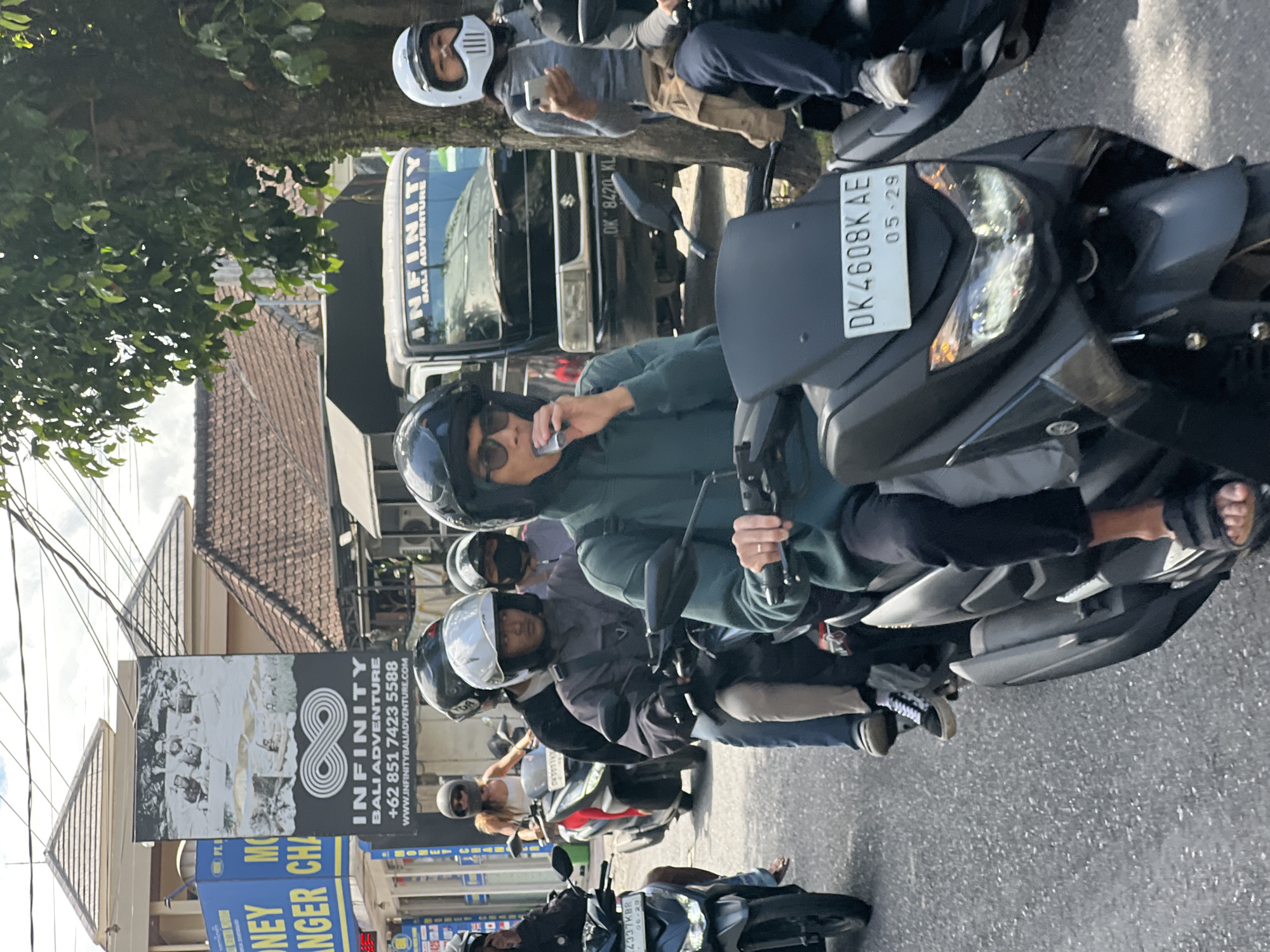 A medium shot captures a traffic scene on a bright day in Ubud, Indonesia, as evidenced by a sign for "INFINITY BALI ADVENTURE" and a license plate reading "DK 4608 K AE". Several individuals are riding motorcycles in a procession. In the foreground, a person wearing a dark green hooded sweatshirt and sunglasses is riding a black motorcycle, holding what appears to be a cigarette or vape pen. Behind them, a person in a dark jacket and light-colored pants is also on a motorcycle. To the right, a person with a white helmet is holding a smartphone while riding. In the background, a black truck with "INFINITY BALI ADVENTURE" written on it is visible, along with other motorcycles and buildings lining the street. The overall environment suggests a bustling urban setting with a clear sky.