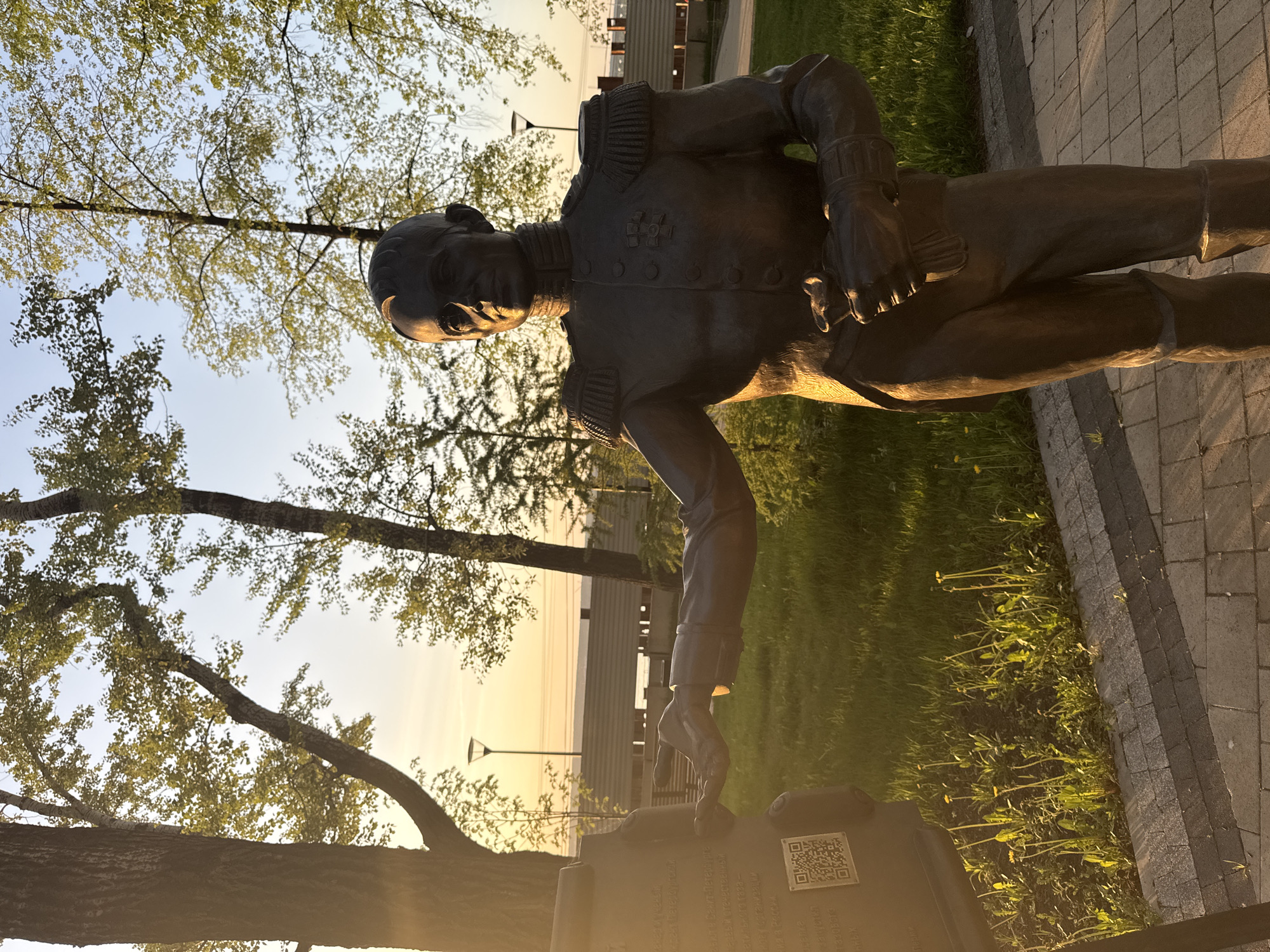 A bronze statue of a man in a military uniform stands on a stone pedestal. The man is depicted with a stern expression and is wearing a long coat with epaulettes. His left hand rests on the pedestal, while his right hand is tucked into his jacket. The pedestal has a plaque with text and a QR code. The statue is located in a park-like setting in Perm, Russia. The background includes trees with leaves, a building in the distance, and a view of a body of water. The time of day appears to be evening, as indicated by the golden light of the setting sun.