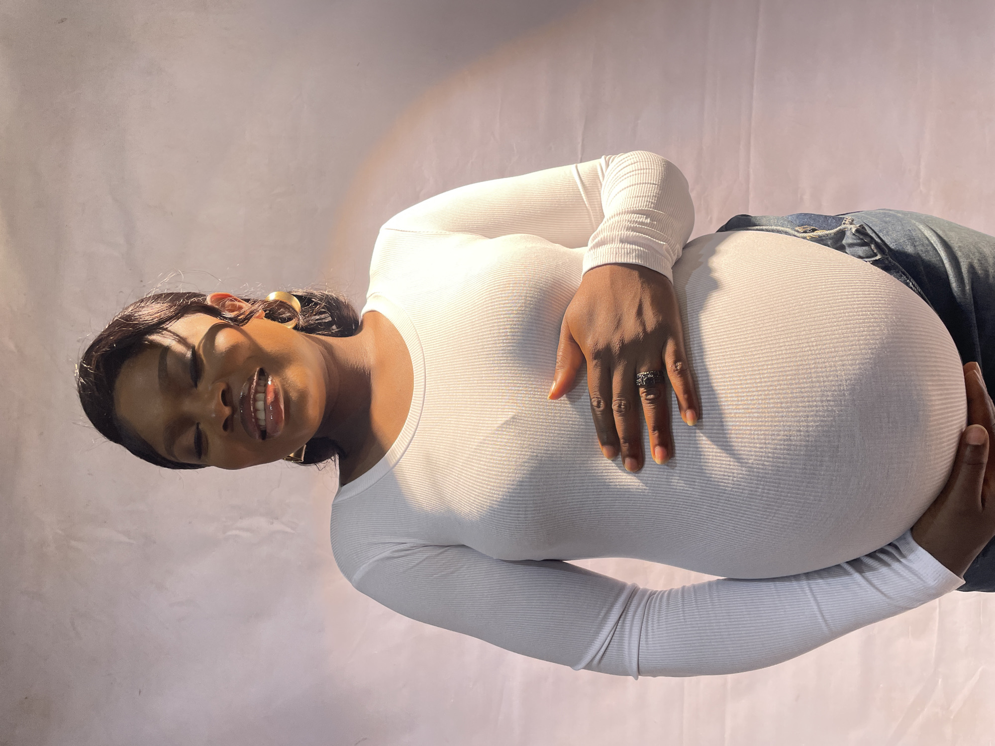 A pregnant woman is posing for a portrait. She is wearing a white long-sleeved shirt and blue jeans. She is holding her baby bump with both hands. She is smiling. The background is a white wall. It's unclear where the photo was taken or if it was taken in Jos, Nigeria.