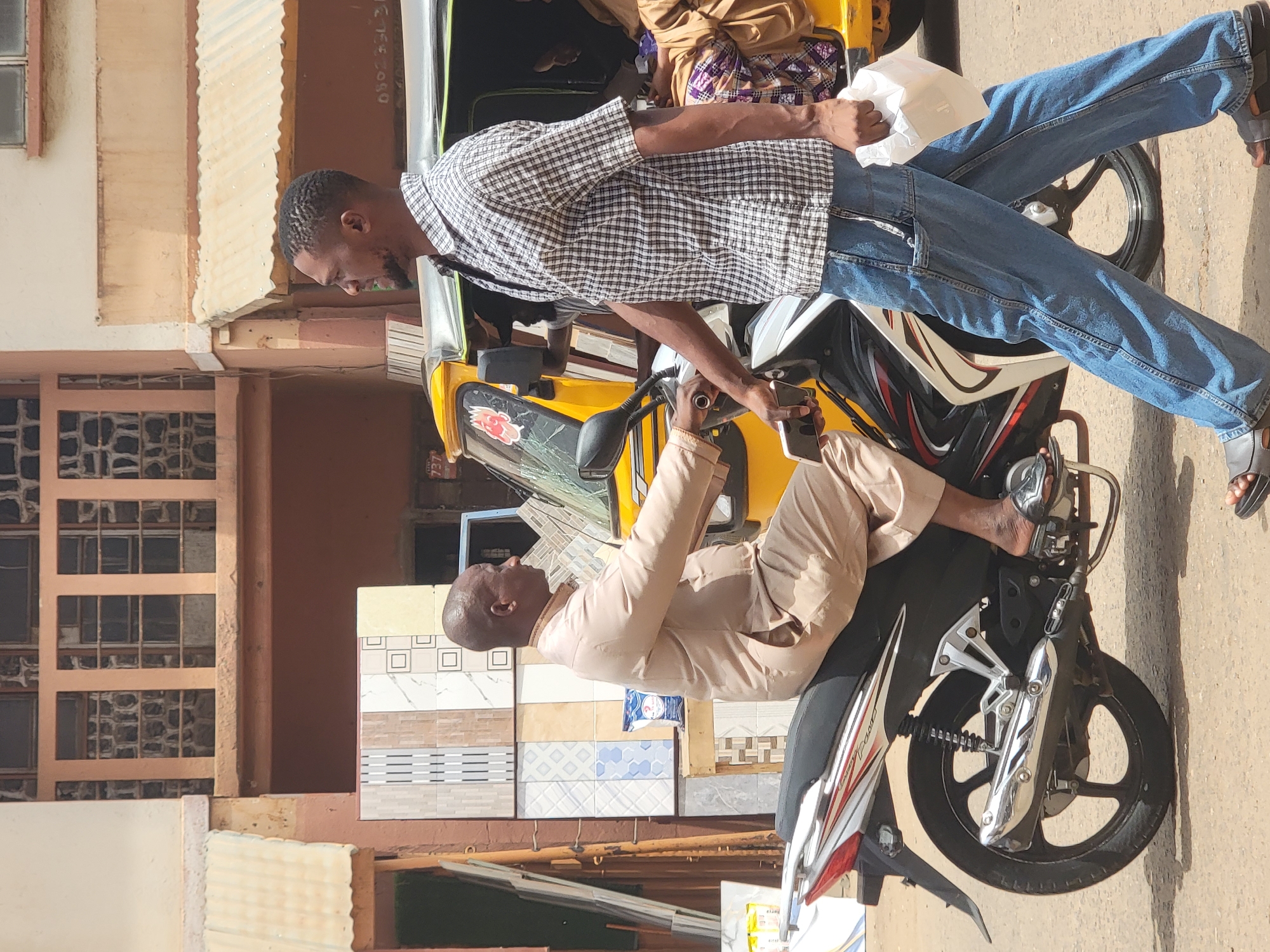In Maiburiji, Nigeria, a man in a checked shirt and jeans is bending down, seemingly conversing with another man sitting on a motorcycle. The man on the motorcycle is wearing a tan suit. A yellow auto-rickshaw is parked behind the motorcycle. Tiles are stacked nearby, suggesting a possible business or construction site. The environment appears to be a street scene.