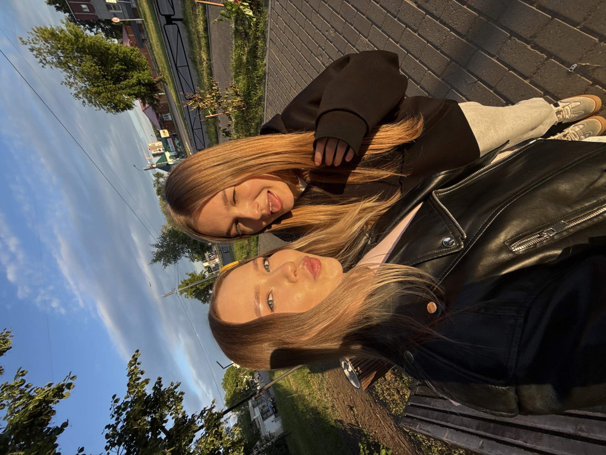 Two young women pose for a selfie outdoors during the golden hour in Chaikovsky, Russia. The woman on the left, with blonde hair and light eyes, wears a black leather jacket over a light-colored top and makes a kissing face towards the camera. The woman on the right, also with blonde hair, wears a black hooded sweatshirt and light grey sweatpants with light-colored sneakers, winking and sticking out her tongue playfully. Her right hand is near her left shoulder, touching her hair.
The scene is an urban street or park area with warm, directional sunlight casting long shadows. The ground consists of light grey interlocking pavers, forming a sidewalk. To the left, a wooden park bench is visible. A low black metal barrier separates the sidewalk from a grassy strip and a paved road beyond. Trees with green foliage are present throughout the background, indicating summer or early autumn.
In the mid-distance on the right, several buildings are visible, including a red-roofed structure and a distinctive green-roofed building with white walls. A sign with Cyrillic text, partially obscured but appearing to be "Дом апте...", suggesting a "Pharmacy," is visible above the entrance of the green-roofed building. A red circular sign with a white horizontal bar, indicating "No Entry," is also visible near the red-roofed building. In the far distance, a construction crane rises above the skyline. The sky is blue with elongated, light-colored clouds. Overhead power lines and streetlights are also present.