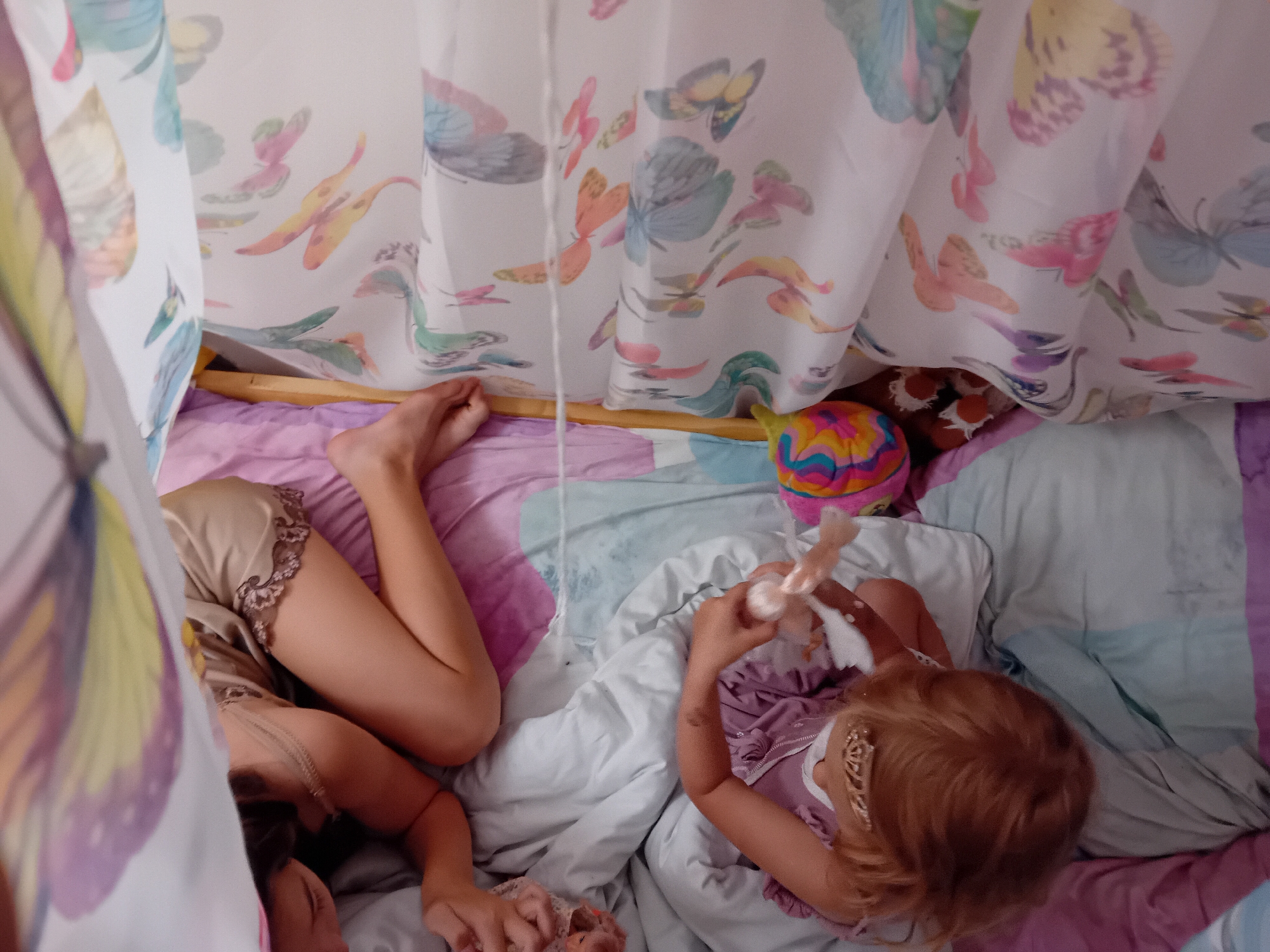 The image depicts two young girls inside a canopy bed. The canopy is decorated with a pattern of colorful butterflies and feathers. One girl, on the right, with blonde hair and wearing a purple top, is holding a doll and playing with it. The other girl, on the left, is lying down with her legs extended and is wearing a beige sleep dress. The bed is covered with pink and purple sheets, and a colorful ball is visible in the background. The scene appears to be an indoor setting, possibly a bedroom in a home. The image does not contain any explicit information about the location in Chuhuiv, Ukraine.