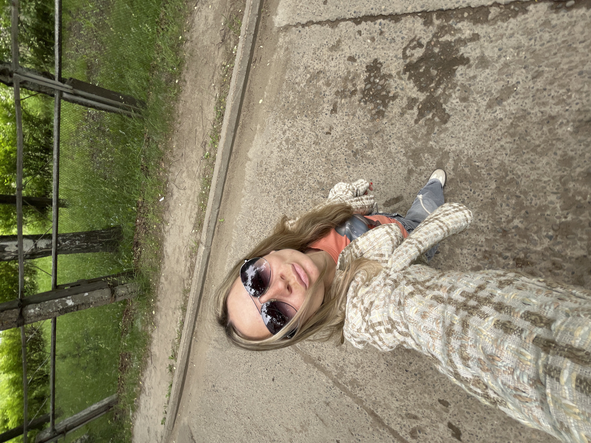 A blonde woman with light skin, wearing sunglasses, an orange shirt, a beige plaid jacket, and jeans, is taking a selfie outdoors on a concrete path. She is standing near a fence and tall green grass. The scene appears to be in a park or outdoor recreational area in Chaikovsky, Russia. The woman is looking towards the camera, with her lips pursed. The lighting suggests it is daytime.