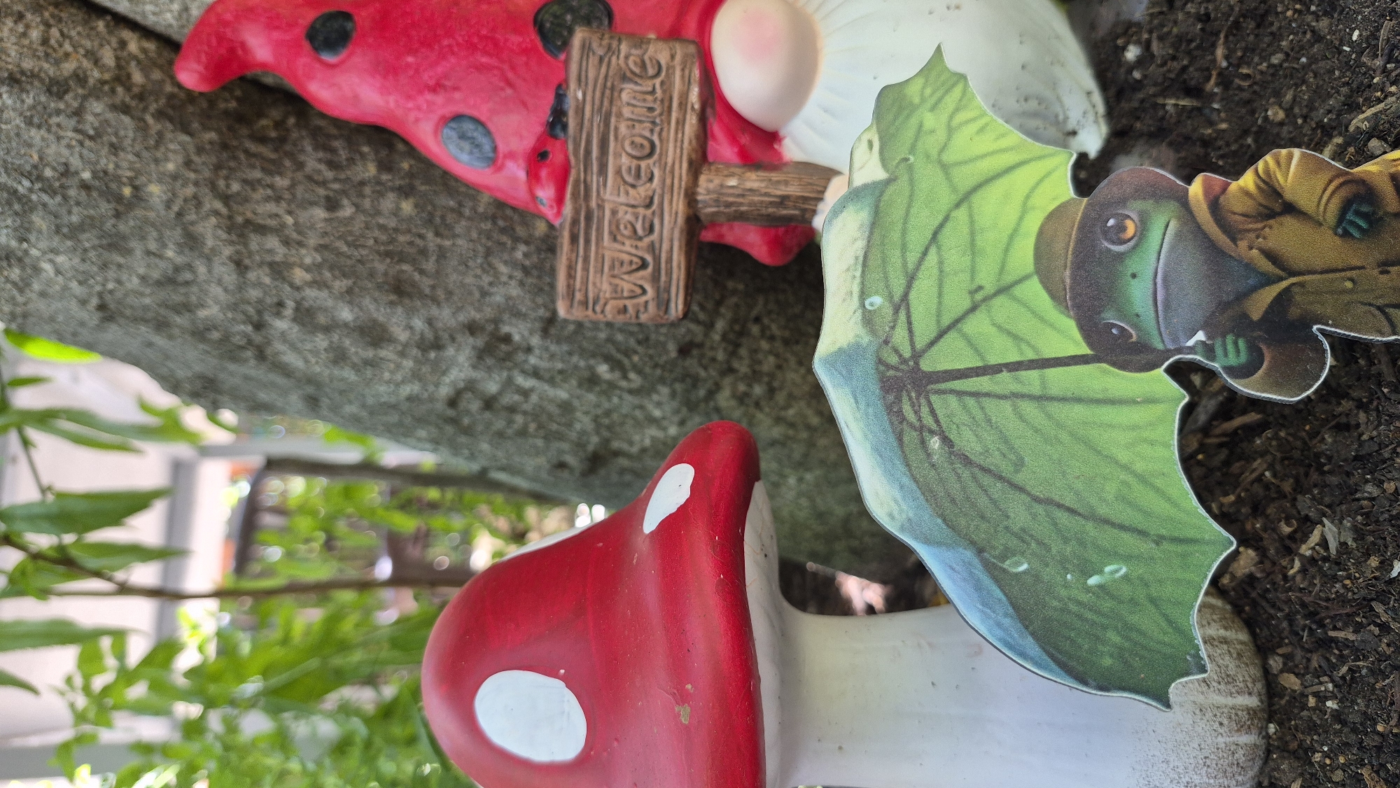 An outdoor, eye-level, medium shot captures a garden scene with a gnome and a mushroom. A large, red mushroom with white spots stands on the left side of the frame. To the right of the mushroom, a gnome with a red hat and black spots is partially visible, holding a small wooden sign that reads "Welcome". In the foreground, a cutout of a frog wearing a yellow raincoat and a hat holds a green umbrella over its head. The background shows a tree trunk and blurred green foliage. The scene is set on dark soil, suggesting a garden or park environment.
