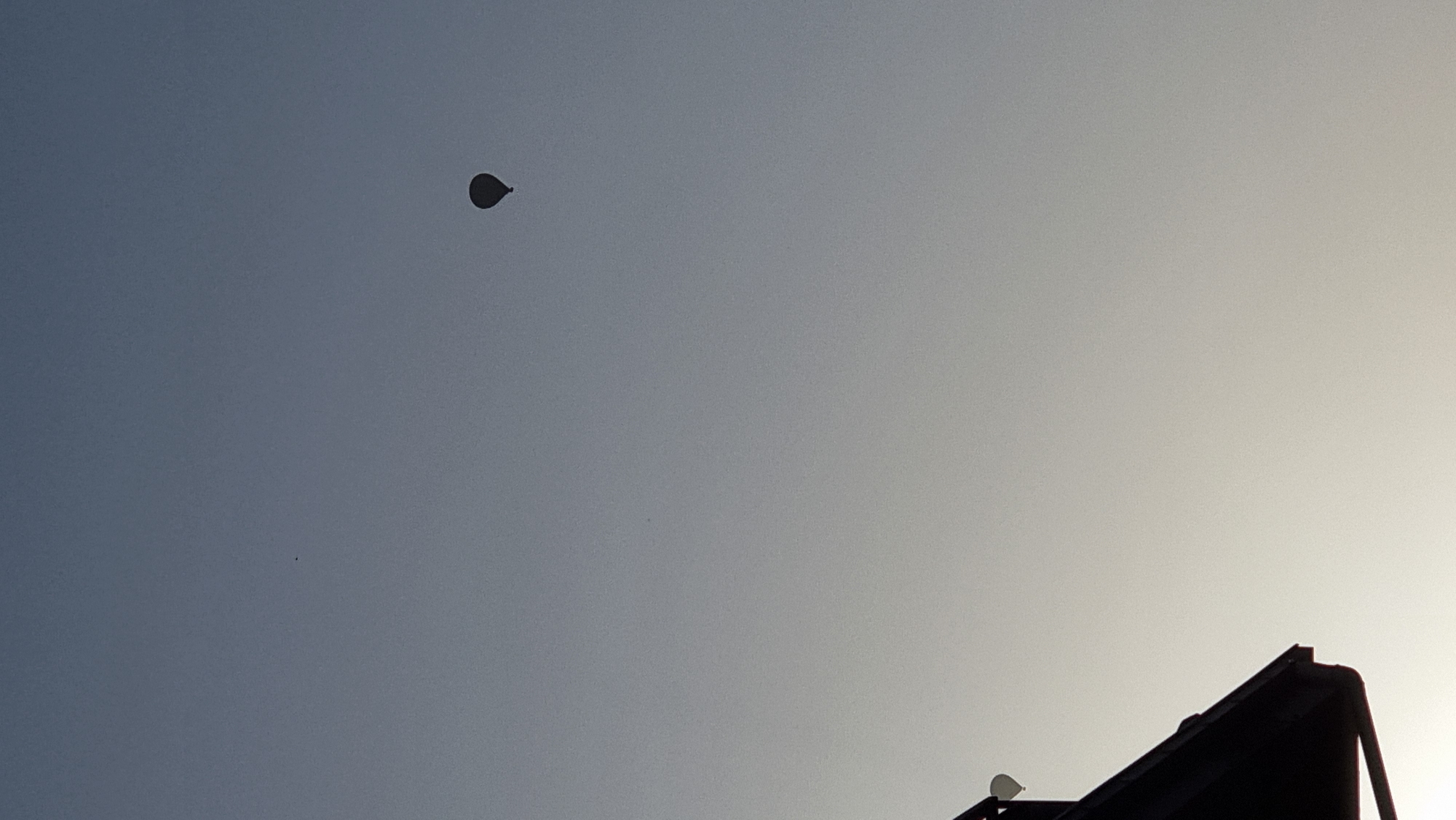 A lone hot air balloon is visible in the sky above Sant Joan Les Fonts, Spain. The balloon is dark and appears to be a silhouette against the pale, gradient sky. In the foreground, a dark, silhouetted section of a building's roof is visible on the left side of the image. The building appears to have a gutter and a portion of a white structure attached to it, possibly a light fixture or part of the roof. The overall scene suggests an early morning or late evening setting due to the soft lighting and the presence of the hot air balloon, which is often seen during these times.