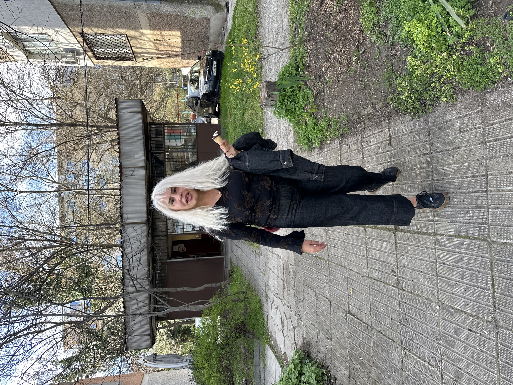 A woman with long white hair is standing on a sidewalk in Sofia, Bulgaria. She is wearing a black blouse, dark denim jacket, and dark denim pants, and black sneakers. She appears to be holding an ice cream cone in her left hand. The sidewalk is made of gray paving stones. To her right is a grassy area with some greenery and a tree stump. Behind her is a small building with a corrugated metal roof and bare trees in the background. Further in the background is a black car and a residential building.