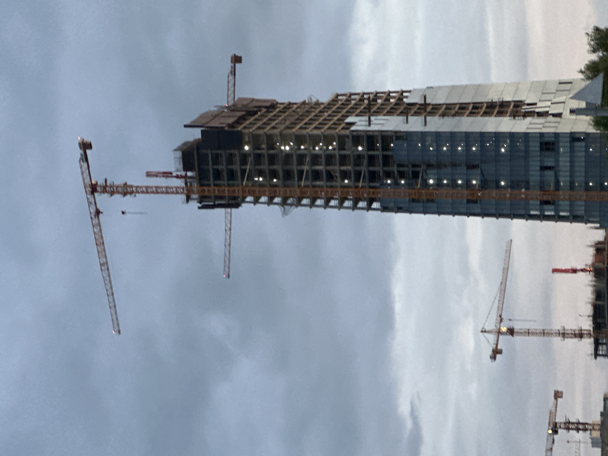 The image depicts a tall, multi-story building under active construction in an urban environment, dominated by several tower cranes against an overcast, twilight sky. The central structure, identifiable as the Magnus Tower (also known as Falcata or BC Pirs) in Minsk, Belarus, features a partially completed facade. The left and lower sections are clad with reflective glass panels exhibiting a modern, angular design. The right and upper sections expose a concrete and steel framework, with multiple internal lights illuminating various floors.
Five tower cranes are present. A prominent, very tall orange and yellow tower crane stands immediately adjacent to the building, its long jib and counter-jib extending over the structure and displaying illuminated red anti-collision lights. A second crane's jib with a red light is visible in the upper left of the frame. A third crane's jib with a red light extends from the right side of the main building. A fourth crane, smaller in stature, is visible to the lower left, also displaying red lights. A fifth, smaller red crane structure is partially visible behind another, less defined structure in the lower left foreground. The sky is uniformly cloudy in varying shades of grey and light blue. No individuals are discernible, nor are any specific actions taking place beyond the implied construction activity. Below the main construction, minimal details of other structures and green foliage are visible at the bottom of the frame.