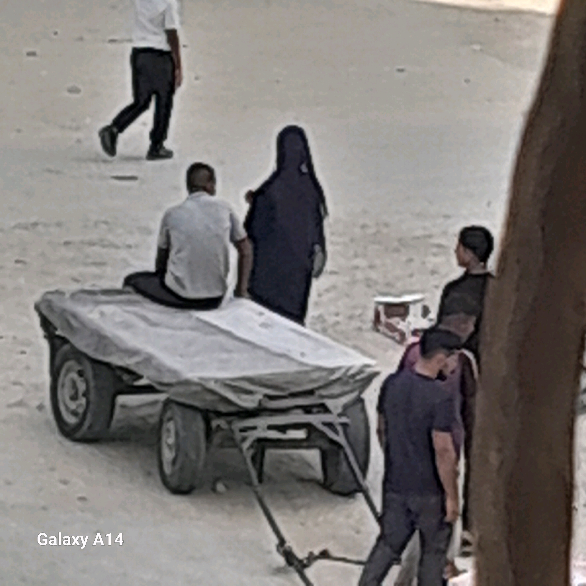 The image depicts a sandy area, potentially a street or open space, in Khan Yunis, Palestinian Territories. Several individuals are present. In the foreground, a cart covered with a light-colored tarp is visible, with its wheels and frame exposed. A person is seated on the cart, facing away from the camera, wearing a light-colored shirt and dark pants. To the left of the cart, a person in dark pants and a white shirt is walking away. Behind the cart, two figures are standing. One is wearing a dark, hooded garment, and the other is facing away from the camera, wearing a dark shirt and pants. Another individual is seen from the back on the right side of the image, leaning against a textured surface, wearing a dark t-shirt and dark pants. A portion of a box with red and white markings is also visible on the ground near the standing figures. The overall scene suggests an outdoor activity or market setting in a sandy environment.