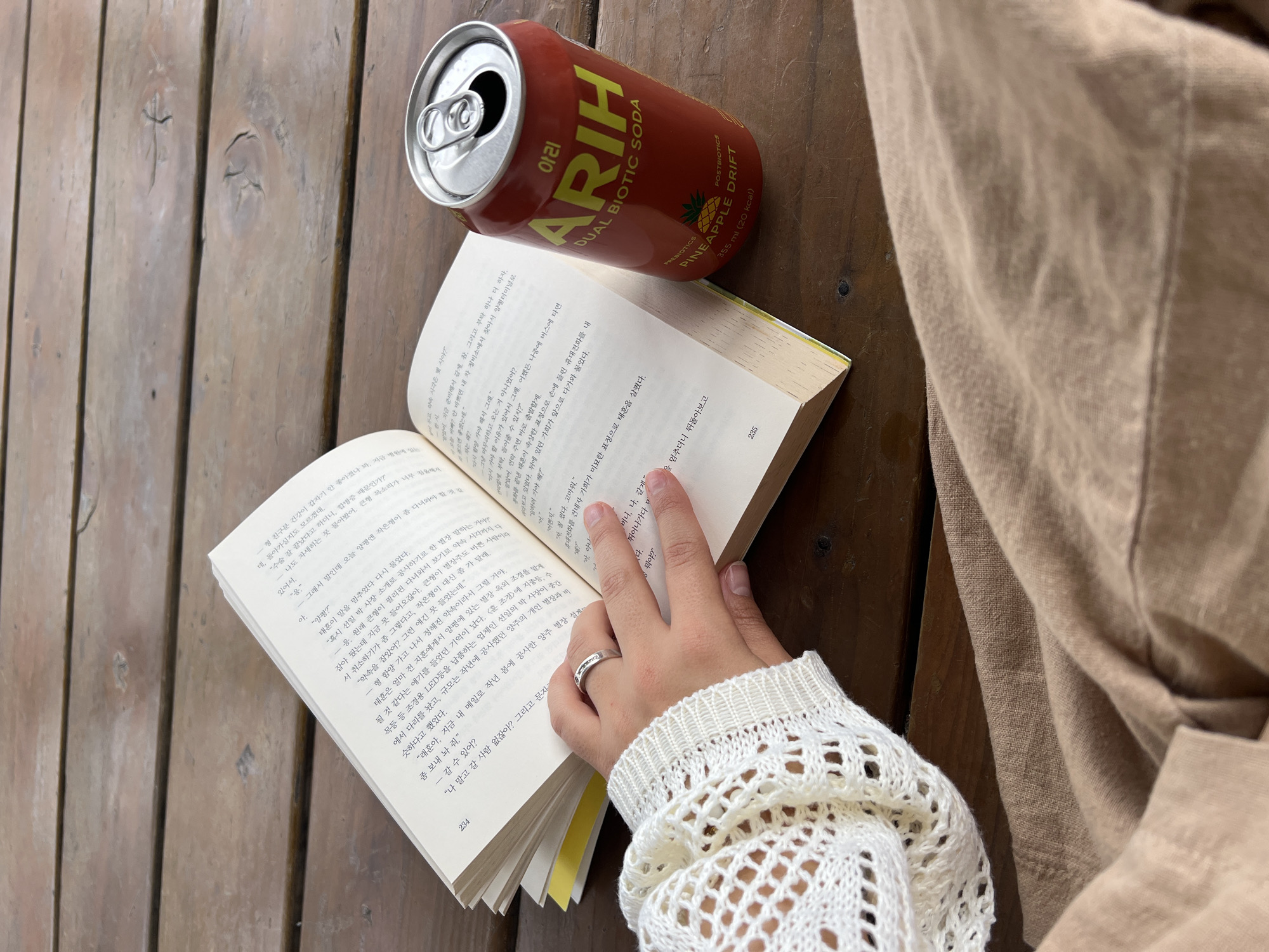 The image depicts a close-up, overhead view of a person's hands holding an open book and a can of "ARIH DUAL BIOTIC SODA" with a pineapple design. The scene is set on a rustic wooden table, suggesting an outdoor or casual setting. The book appears to be a novel, with Korean text visible on its pages. The can is red and metallic, with prominent branding. A person's arm, wearing a white, crocheted sleeve, is visible on the left side of the frame, with their fingers gently holding the book open. The lower portion of the image shows a person wearing a light brown garment, possibly pants or a skirt. The lighting suggests daylight, casting soft shadows. There is no discernible indication of the city of Daegu, South Korea, within the visual elements of the image.