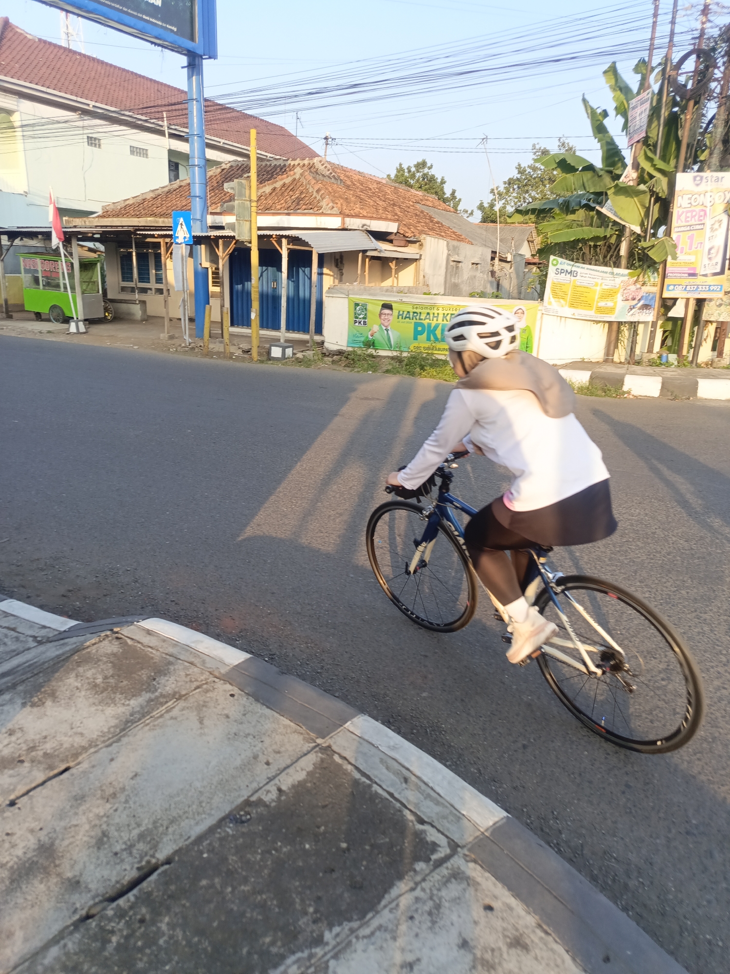 An individual wearing a white helmet, a long-sleeved white shirt, and dark leggings is riding a blue and white road bicycle on a paved road. The cyclist is seen from the rear and right side, with their right leg extended towards a pedal. The bicycle has thin wheels with visible spokes. In the background, there are buildings with reddish-brown tiled roofs, a green food cart with a white awning and wheels, a traffic signal with a blue sign depicting a white pedestrian crossing, and various posters and banners attached to poles and walls. One banner features an image of a man in a green suit and the text "SELAMAT & SUKSES HARLAH KE-XX PKB". Another poster has the text "star NEONBOX KEREN". Banana trees and other vegetation are also visible. The scene appears to be in a semi-urban or town environment, consistent with a location in Desa Gunungsimping, Indonesia, given the cultural context of the banners and architecture. The lighting suggests it is either early morning or late afternoon, with long shadows cast on the road. A concrete curb and sidewalk are visible in the foreground.