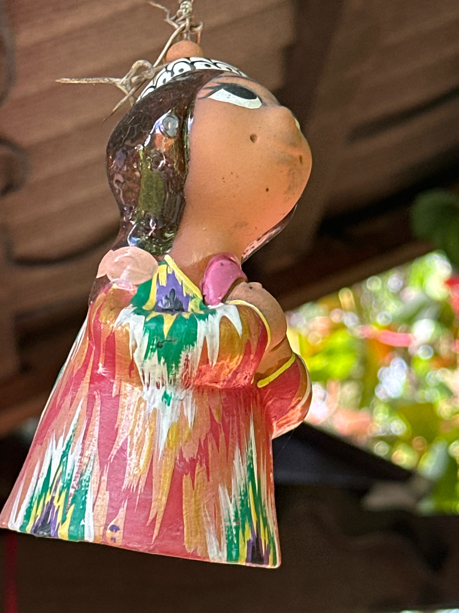 An artistic, close-up, eye-level shot shows a colorful, ceramic figurine of a person hanging from a string. The figurine is adorned with bright, abstract patterns in shades of red, pink, yellow, green, and white, resembling a dress. The figurine's head is tilted back, with dark, glossy hair, a distinct white headdress with black circular accents, and large eyes. A light pink detail, possibly representing a flower, is visible near the figurine's shoulder. The background is softly blurred, revealing glimpses of green foliage and a dark, wooden structure, suggesting an outdoor or semi-outdoor setting. The overall impression is one of folk art or handicraft, possibly displayed in a garden or courtyard. This image does not provide any specific information about the city of Калодзішчанскі сельскі Савет, Belarus, as it focuses solely on the ceramic figurine and its immediate surroundings.