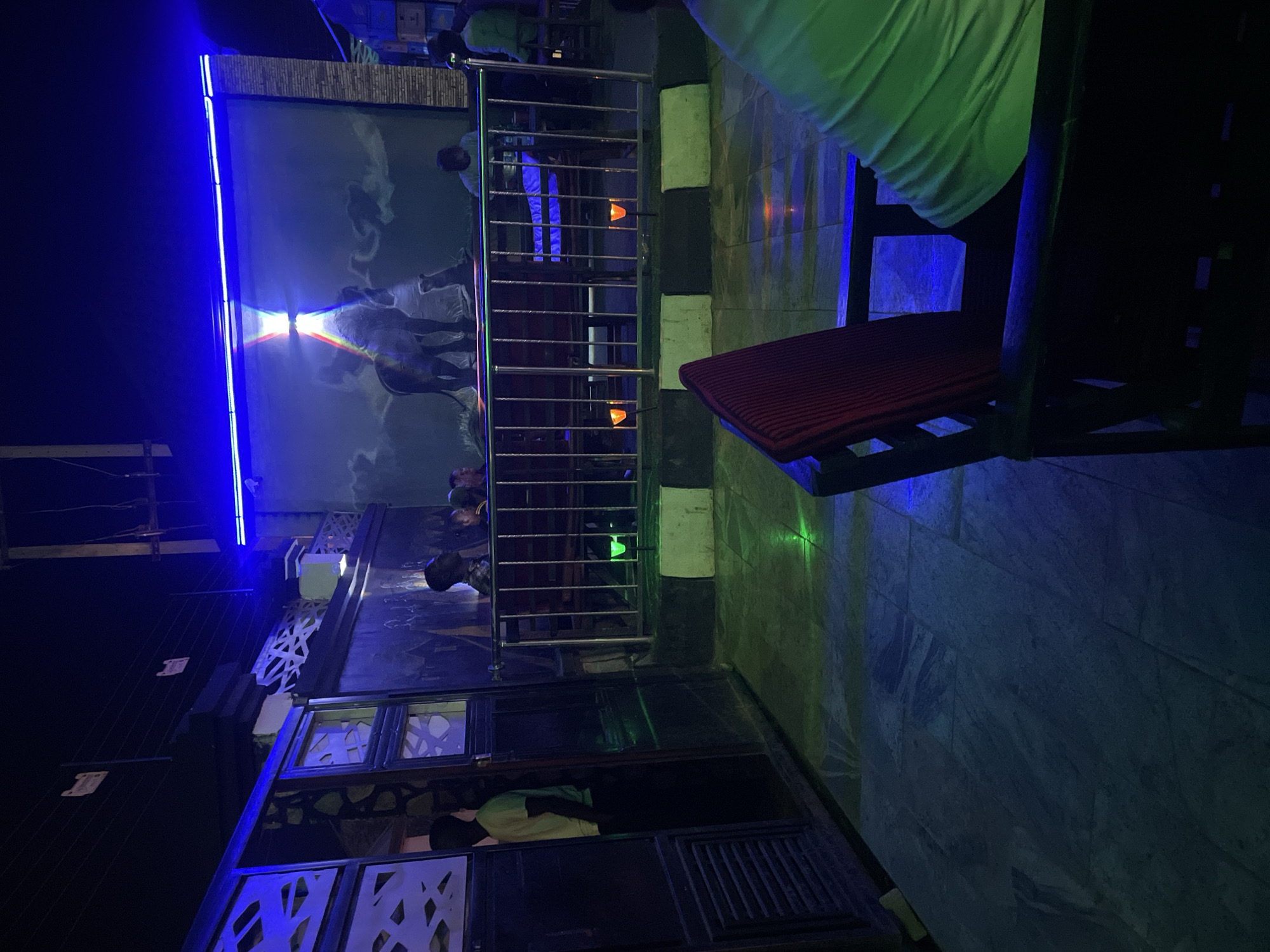 The image depicts an outdoor bar or lounge area in Tumfure, Nigeria, at night. The area is illuminated with blue and green lighting. A mural of horses is visible on a wall with a blue neon light strip. A metal railing separates the upper and lower levels of the lounge. People are present on both levels, their faces mostly obscured. A chair with a red cushion is positioned in the foreground. The floor is tiled with a gray and white pattern. Overall, the scene suggests a modern, dimly lit nightlife venue.