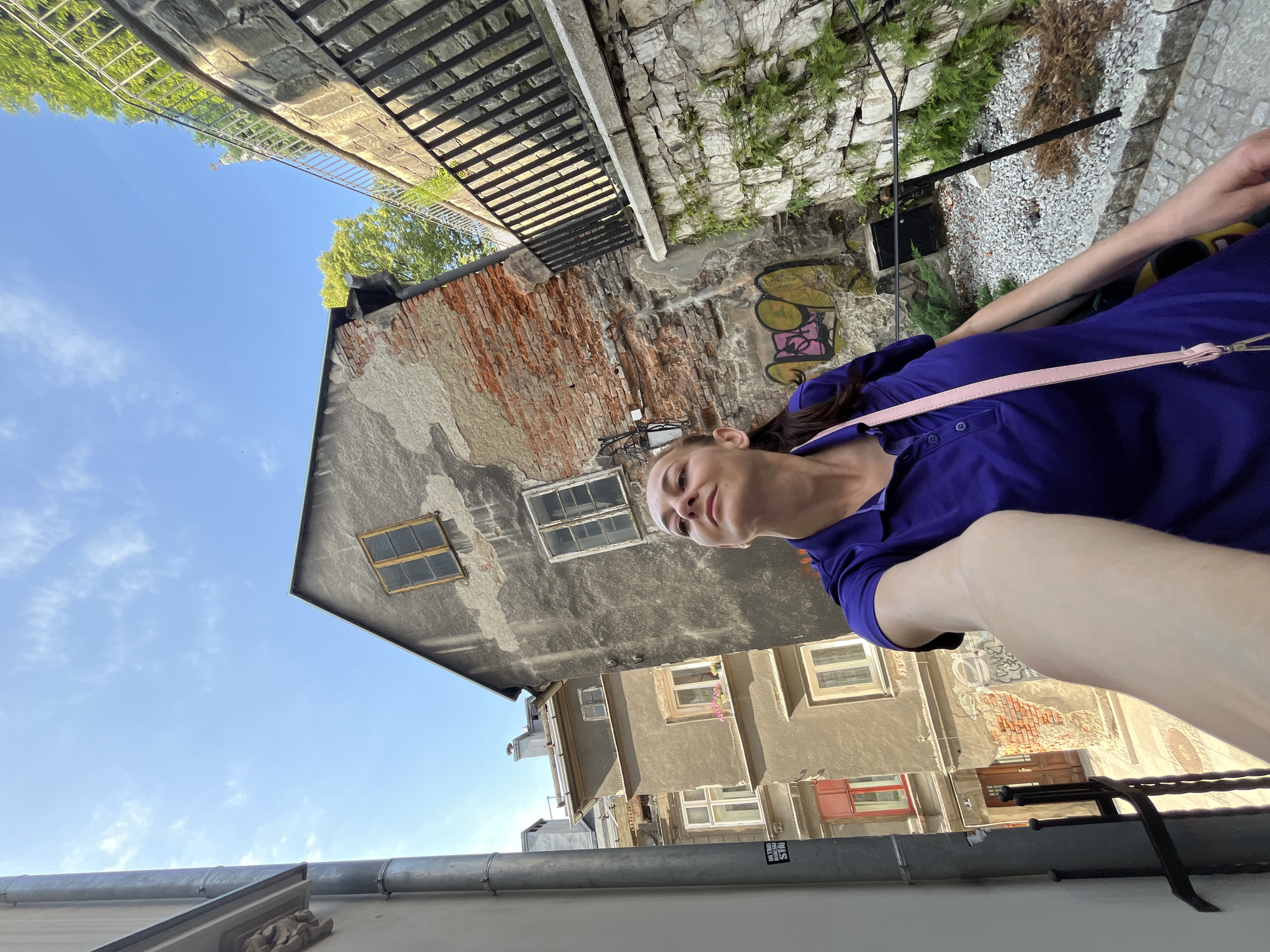 In an urban environment, a woman with her hair pulled back poses for a selfie in a narrow alley. She is wearing a vibrant purple polo shirt and a light pink crossbody bag. The alley's walls are weathered and textured, with exposed brick and patches of peeling plaster. Graffiti adorns one section of the wall. Above, the sky is a clear blue with scattered white clouds. To the right, a stone wall topped with a metal railing rises, and lush green vegetation grows along it. In the background, a multi-story building with several windows can be seen. The ground is paved with cobblestones.