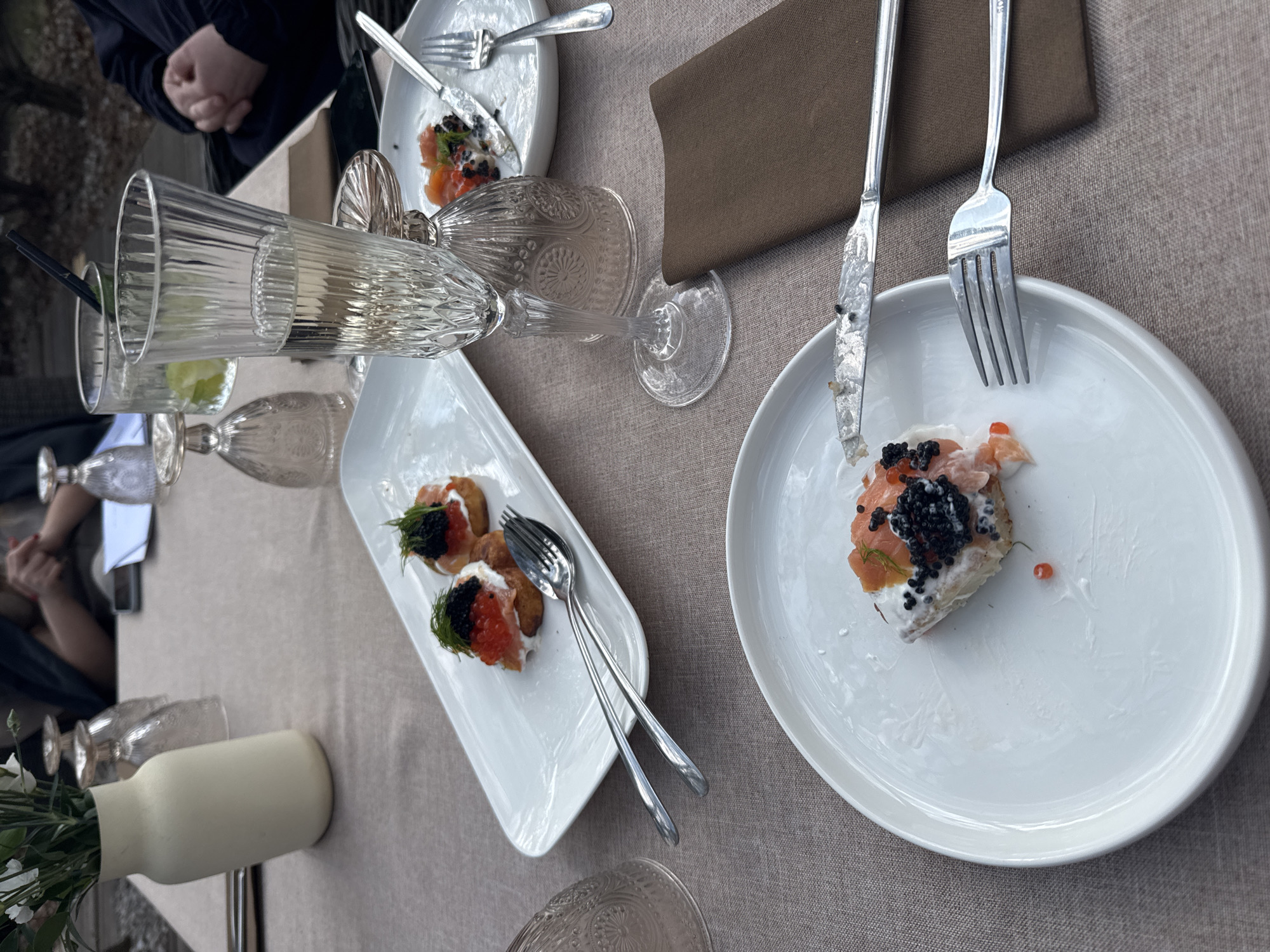 The image depicts a dining table set for a meal. Plates are arranged with partially eaten appetizers featuring salmon and caviar. Cutlery is laid out beside the plates. Several glasses, some empty, are also on the table. A beige napkin is placed near one of the plates. A small vase with flowers is situated at the edge of the table. The setting suggests a restaurant or private dining event. There is no specific information available in the image to confirm this happened in the city of Konstancin-Jeziorna, Poland.