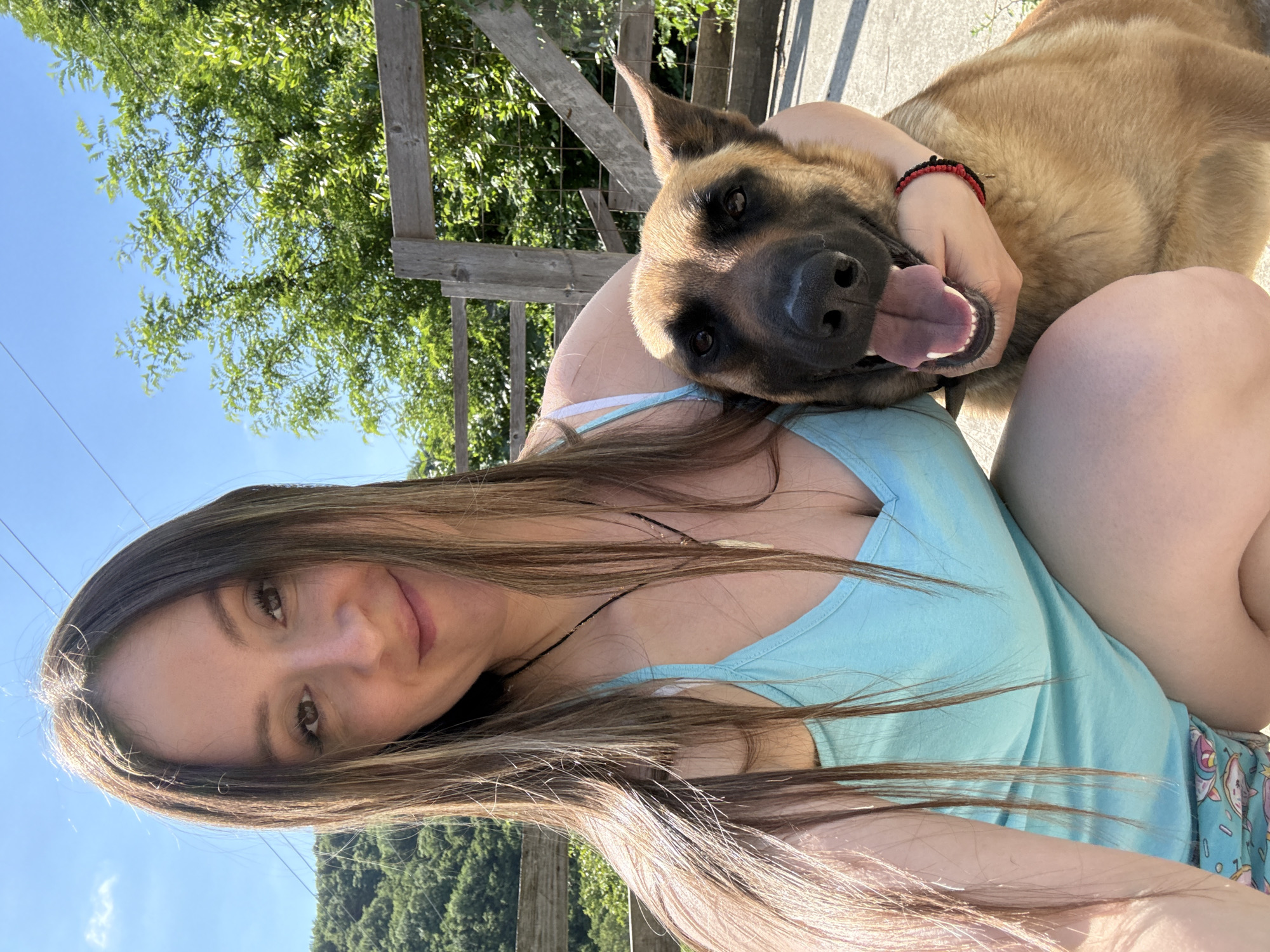 A woman with long brown hair and a light blue tank top is posing for a selfie outdoors. She is embracing a German Shepherd with a black mask and tan fur. The dog is panting with its tongue out. They are positioned in front of a wooden fence and lush green trees under a bright blue sky. The background appears to be a rural or suburban setting, possibly in Kroyanskoye, Russia, given the landscape.