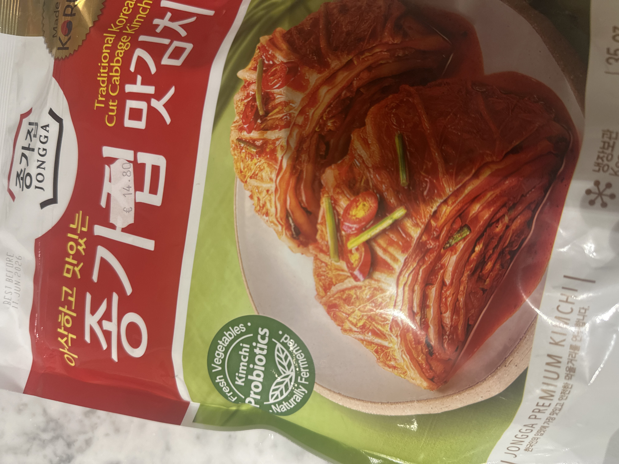 The image captures a close-up, angled view of a Jongga brand kimchi package, likely situated in Milan, Italy, as indicated by the Euro price tag. No people are visible, and the setting appears to be an indoor surface, possibly a countertop, with no specific time of day or weather discernible.
The package is predominantly red and white, featuring a prominent image of ready-to-eat cut cabbage kimchi on a plate, illustrating its vibrant red hue, green accents from spring onions, and chili pieces, suggesting a fresh and spicy flavor.
Key visible text includes the brand "JONGGA" (종가집) and its Korean name, along with the description "Traditional Korean Cut Cabbage Kimchi" and '맛김치'. A circular green emblem highlights "Fresh Vegetables," "Kimchi Probiotics," and "Naturally Fermented." The origin is noted as "Made KOR" (Korea).
A price sticker displays "€ 14.80," and a "BEST BEFORE 11 JUN 2026" date is clearly printed. Other Korean text on the package reads "아삭하고 맛있는 종가집 맛김치" (Crisp and delicious Jongga Kimchi) and "JONGGA PREMIUM KIMCHI | 한국인의 입맛에 가장 맛있고 안전한 먹을거리를 연주합니다." (It offers the most delicious and safe food to the taste of Koreans), alongside "냉장보관" (Refrigerate).