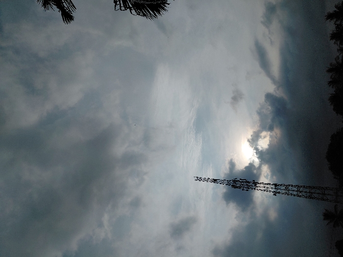 The scene is located in Mns Alue, Indonesia, under an overcast sky. A tall, lattice-structure communication tower stands on the left side of the frame, silhouetted against the sky. The sun is partially visible and diffused by dense, dark gray clouds positioned behind and around it, creating a bright central glow. The sky is predominantly filled with varying shades of gray clouds, some appearing heavy and dark. The lower portion of the image contains dark silhouettes of vegetation, including the tops of trees and distinct palm fronds visible on the far right. No people or specific activities are present in the image.