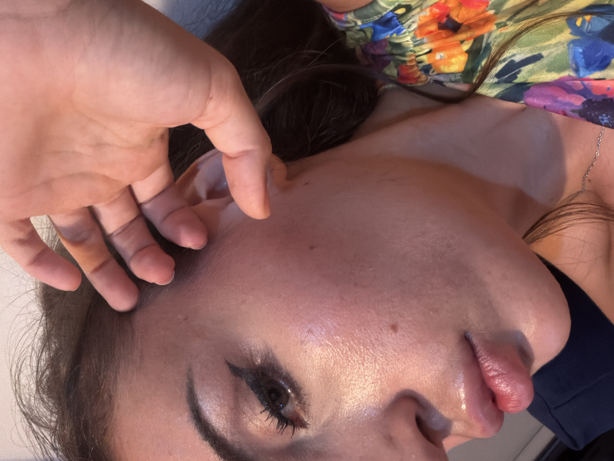 A close-up, profile view of a woman's face and upper body is depicted. The woman has dark hair and is wearing makeup, including eyeliner and mascara. Her right hand is positioned near her face, with her fingers touching her forehead and hair. She is wearing a brightly colored floral print top. The lighting suggests an indoor setting, likely within a residence in Tersefanou, Cyprus. The scene is focused on the woman's features, highlighting her skin texture and makeup.