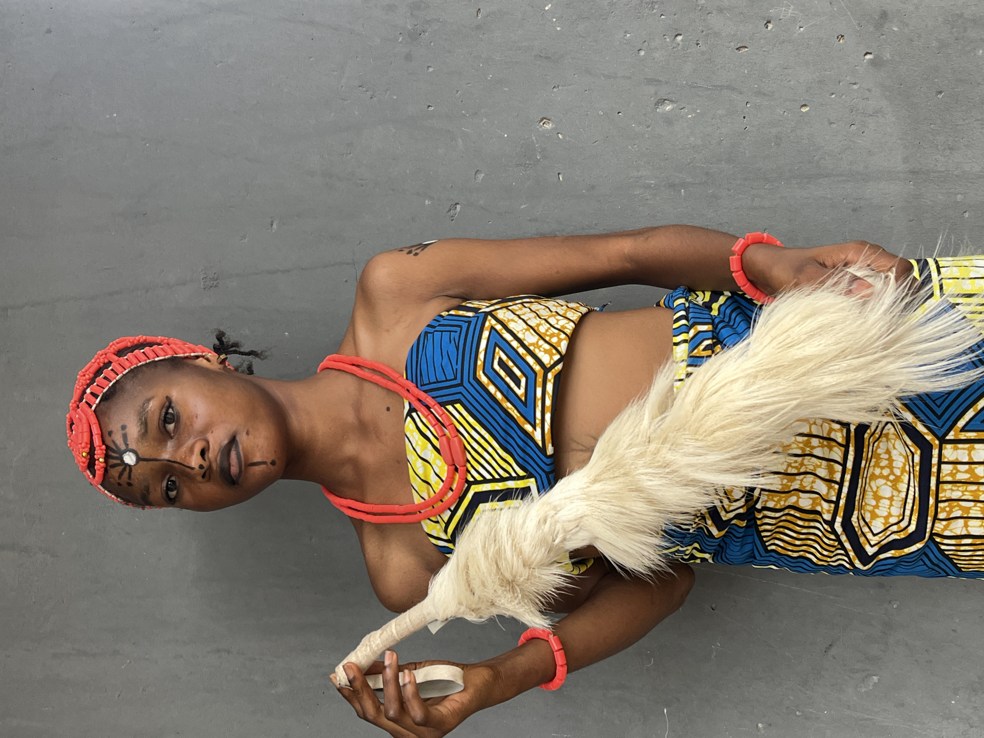 A woman in traditional Nigerian attire stands against a gray background. She wears a beaded red headband, necklace, and bracelet, with tribal markings on her face. Her clothing is a yellow, blue, and black patterned fabric. She holds a white feathered object in her hand. The image is shot indoors with a slightly angled perspective. The location is presumed to be in or related to Badagry, Nigeria based on her attire.