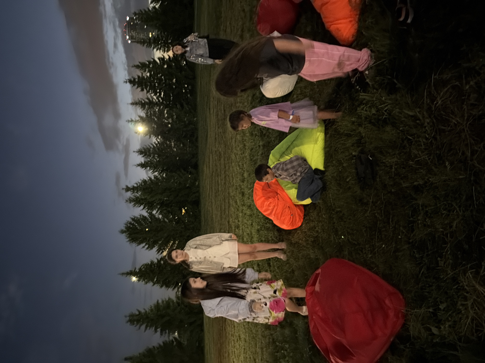 The image shows a group of people, including several children and two adults, gathered in a grassy field at dusk. The sky is dark blue with some clouds. A line of evergreen trees is visible in the background, and a tall building with lights is in the far right background. In the foreground, there are several large, brightly colored beanbag chairs, including red, orange, and lime green. A young girl with long dark hair is standing on the left, wearing a light blue jacket and a floral dress. Another young girl, wearing a white outfit, stands behind her. A boy in a plaid shirt is sitting on the lime green beanbag chair, and a young girl in a purple dress stands next to him. An adult woman in a sparkly jacket is standing on the far right, and another adult woman with long dark hair is in the middle ground, wearing pink pants and a dark top. The people appear to be socializing and enjoying an outdoor activity.